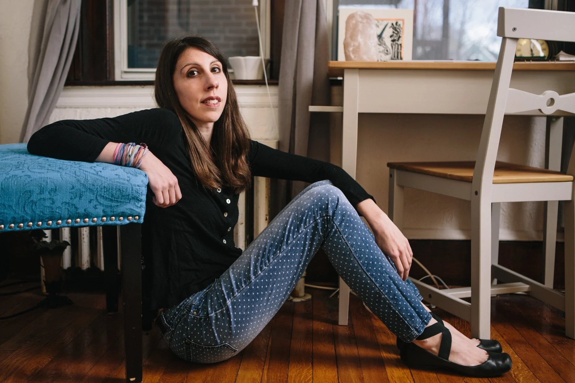 A woman with long brown hair sitting on the floor, leaning against a blue velvet chair, in a room with wooden floors, near a window with curtains, with art and objects on the desk behind her.