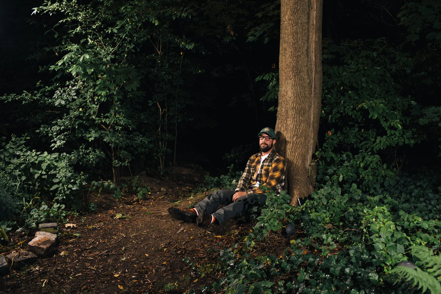 A man with glasses and a beard wearing a plaid shirt sitting against a tree in a dark forest, illuminated by a flashlight.