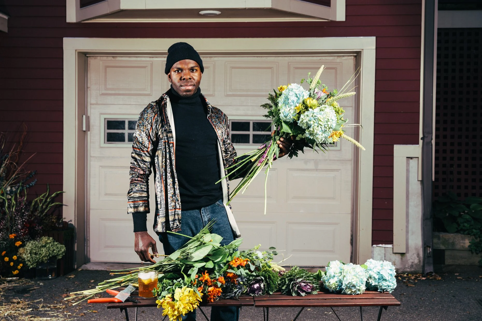 A man in a black beanie and patterned jacket stands in front of a garage door, holding a bouquet of flowers and arranging floral donations on a table outside a house.