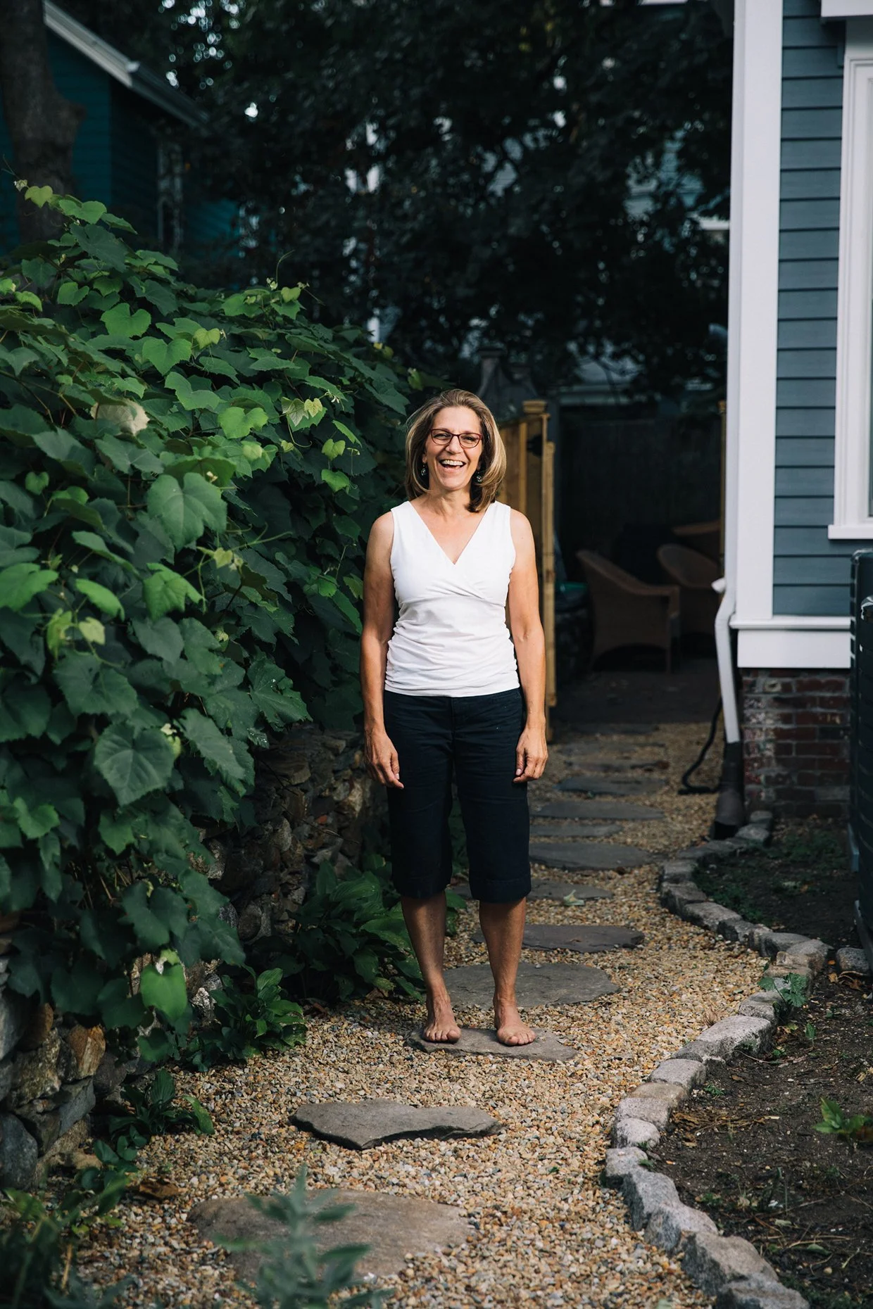 A cheerful woman with glasses and shoulder-length hair, wearing a white sleeveless top and black capri pants, walking barefoot on a stone pathway in a garden yard with lush green bushes and a blue house exterior in the background.