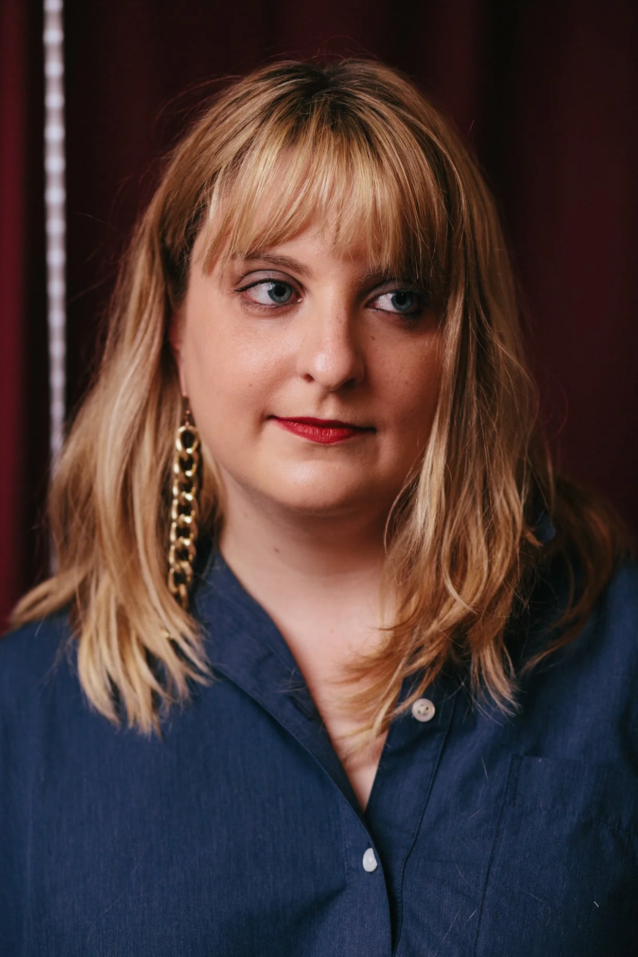 A woman with shoulder-length blonde hair and blue eyes, wearing a dark blue button-up shirt and large gold chain earring, looking slightly to the side.