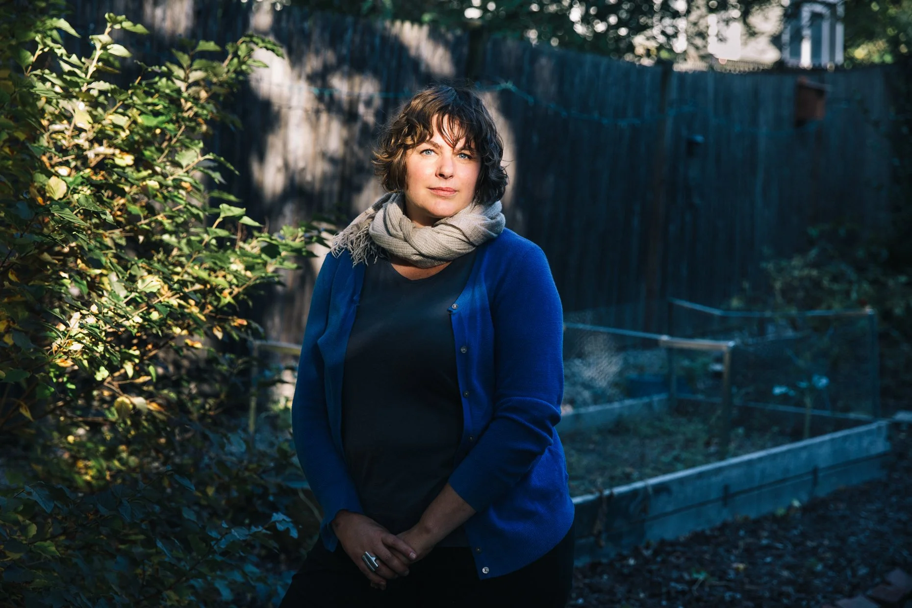 A woman with short wavy brown hair and blue eyes standing in a garden or backyard, wearing a black top, blue cardigan, and a beige scarf, with greenery and a wooden fence in the background.