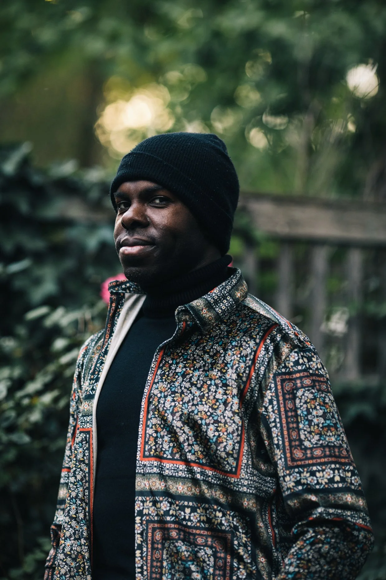 Portrait of a man outdoors wearing a black beanie and a patterned jacket with floral and geometric designs, standing in front of a blurred green background.