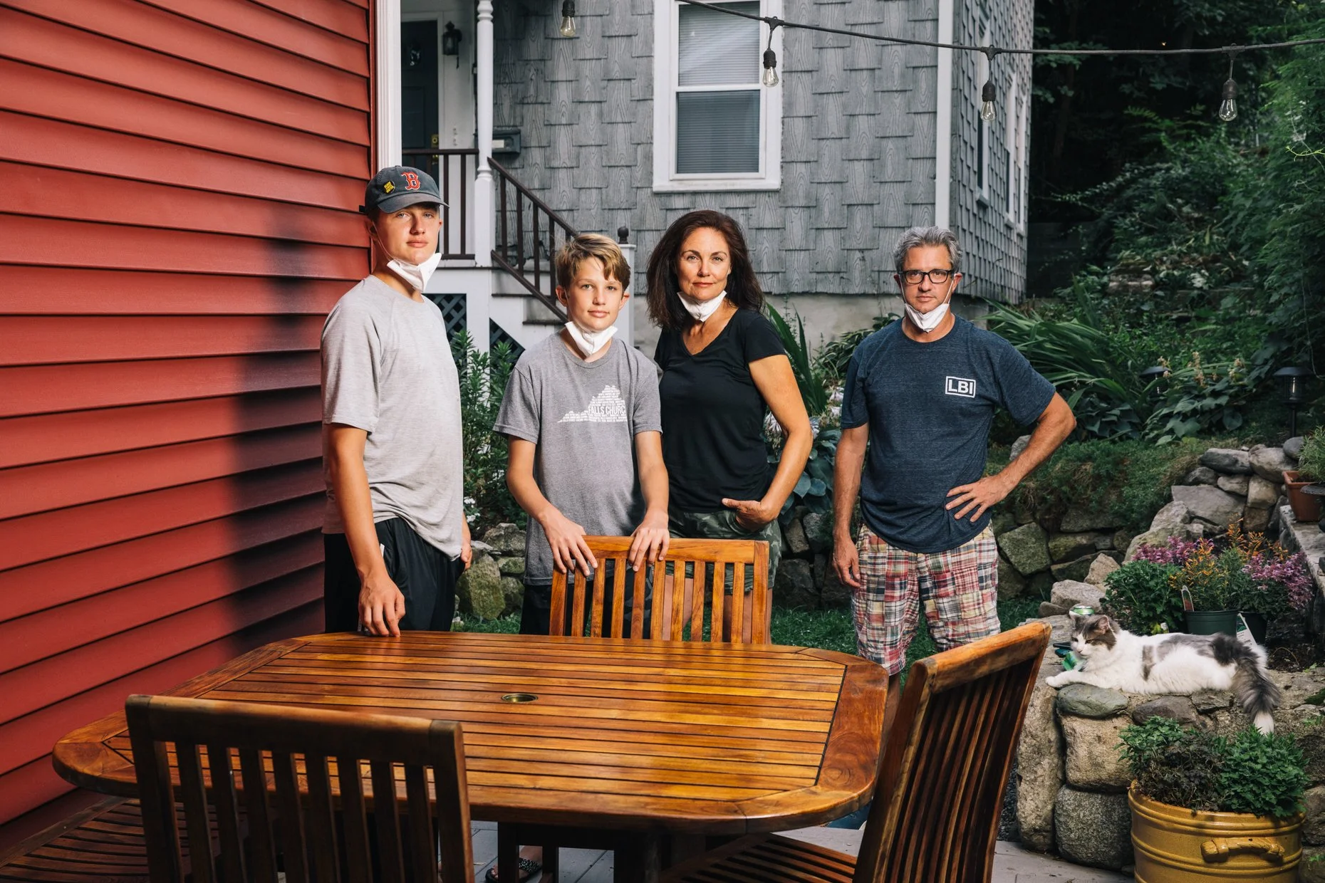 Four people standing outdoors on a patio in front of a house, with a wooden table and chairs in the foreground, and a garden with potted plants, rocks, and greenery in the background. Three of them are wearing face masks pulled down.
