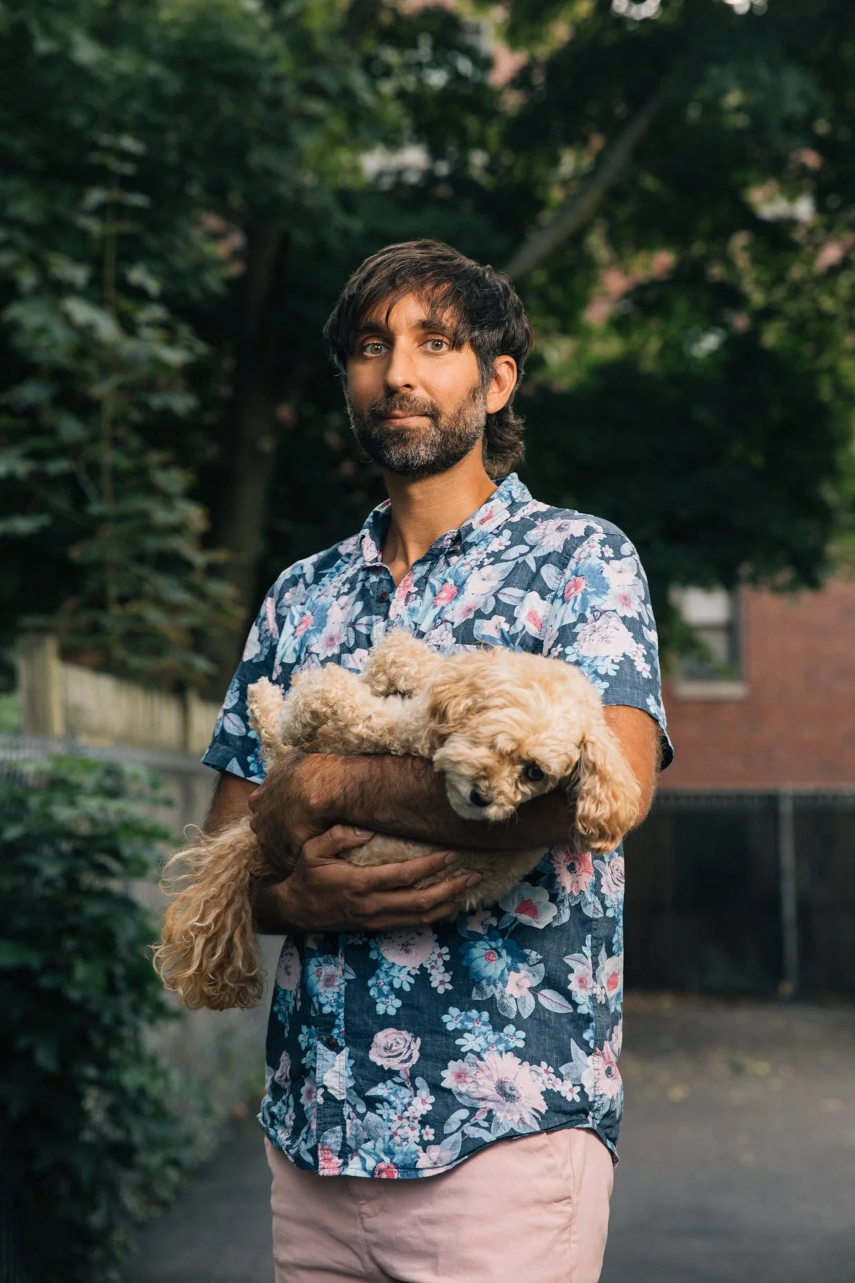 A man with dark hair and a beard holding a light-colored poodle puppy outdoors in a green backyard or park with trees and a fence in the background.