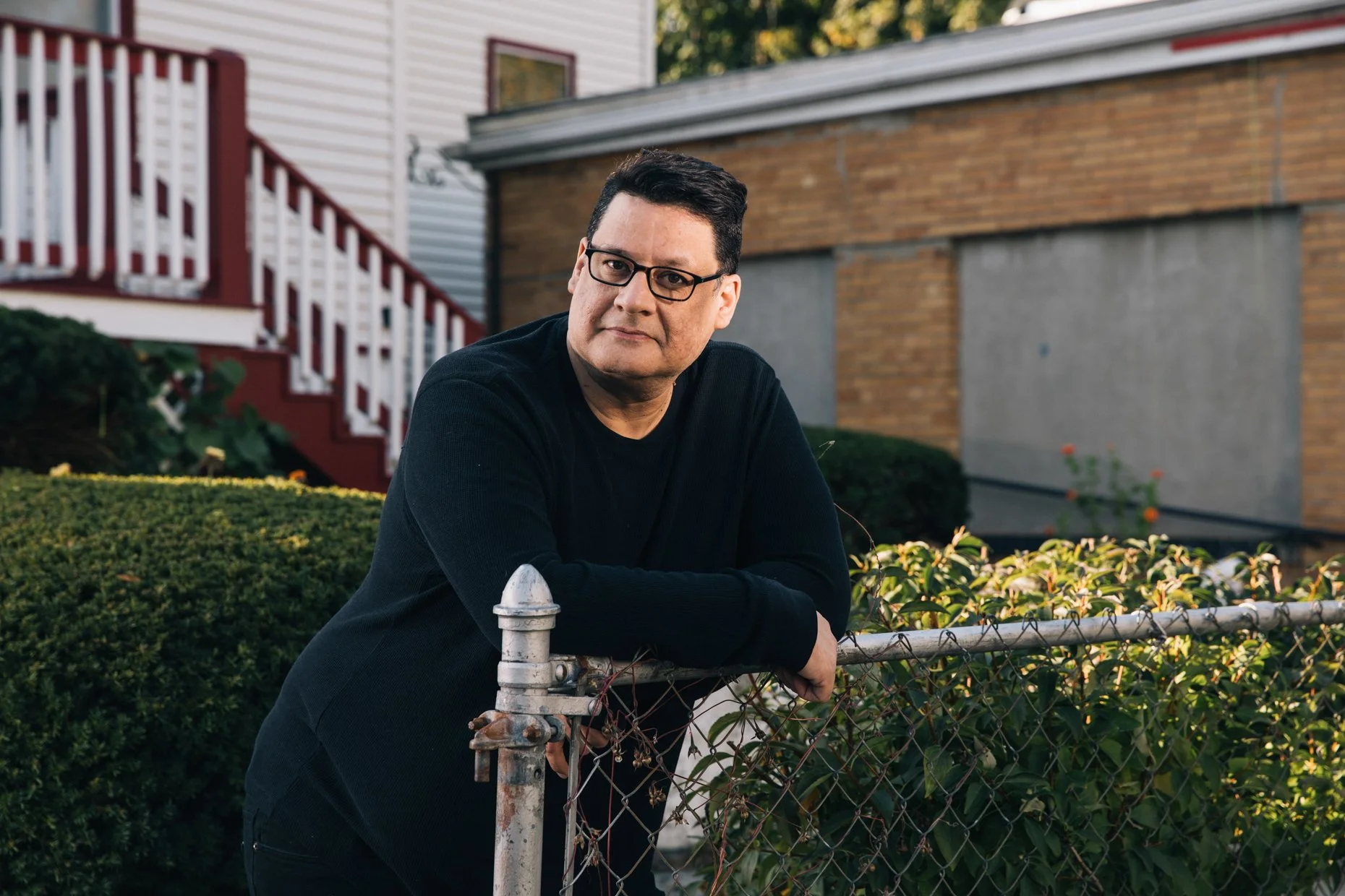 A man with dark hair and glasses leaning on a rusty metal fence in front of a hedge with purple flowers. Behind him is a building with a red staircase, white siding, and brick walls.