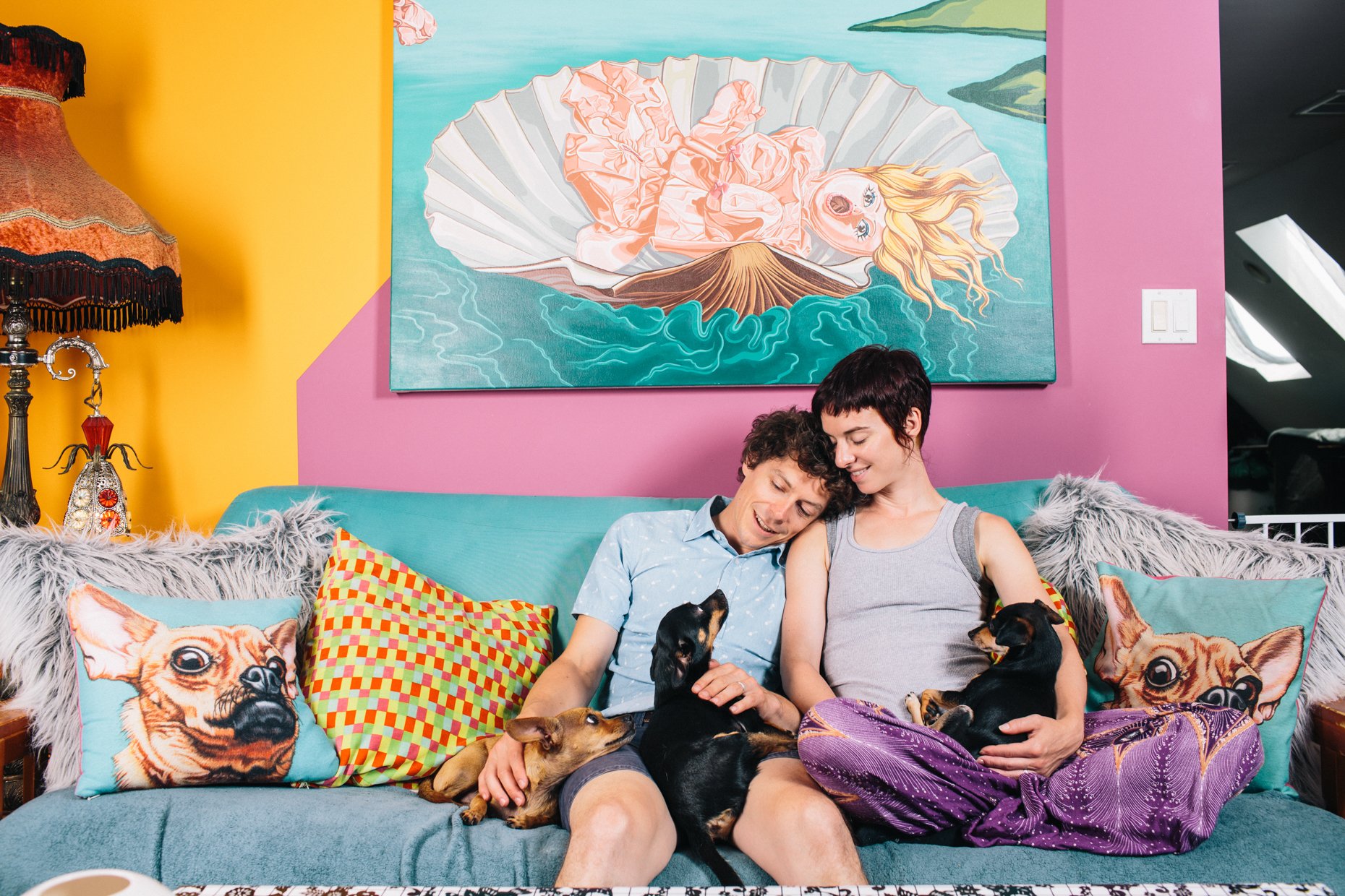 Two people sitting on a blue sofa with colorful pillows, surrounded by several dogs, in a brightly decorated room with yellow and pink walls, a large sea-themed artwork, and a vintage-style lamp.