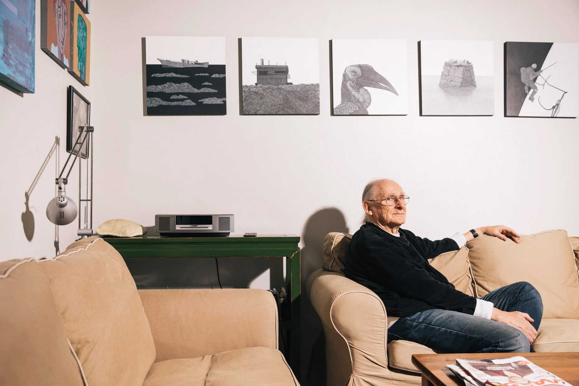 An elderly man sitting on a beige sofa in a living room, holding a remote control, with framed artwork on the wall above him and a green table with a stereo and a pillow beside it.