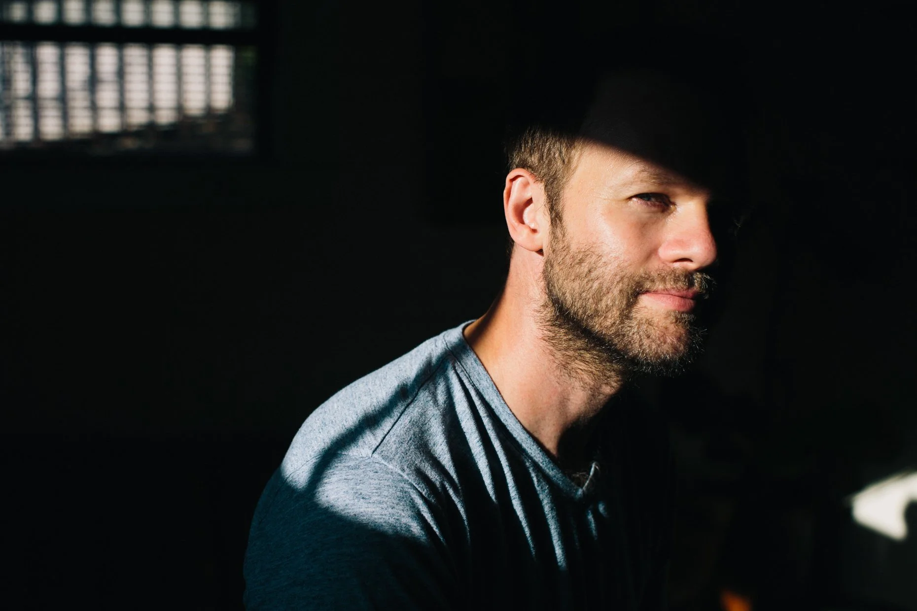A man with short brown hair, a beard, and light skin, wearing a gray t-shirt, is looking to his left with a slight smile. The background is dark with a hint of a window with blinds.