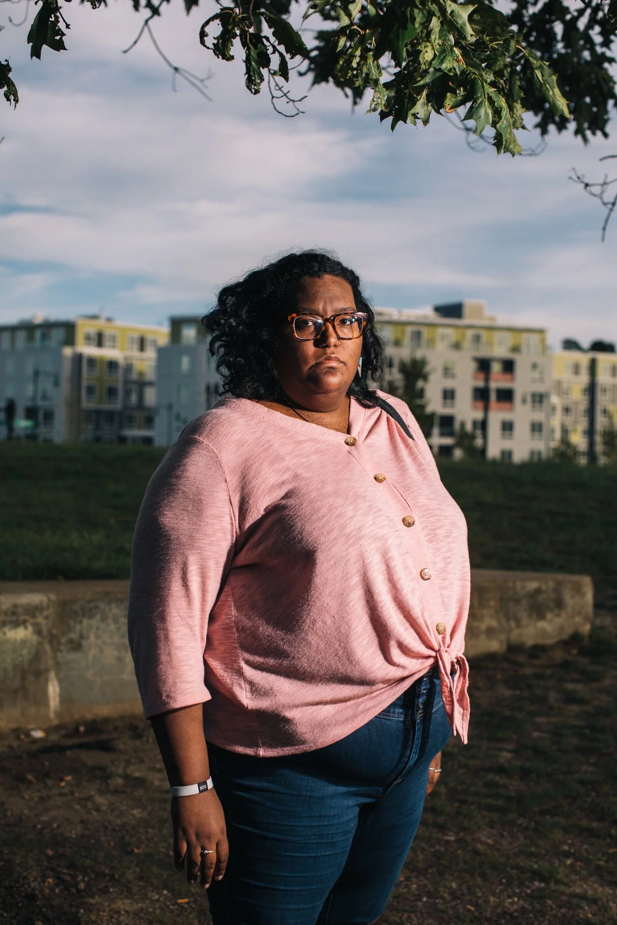A woman with curly black hair, glasses, and a serious expression, wearing a pink button-up shirt and jeans, standing outdoors during sunset with buildings and a tree in the background.