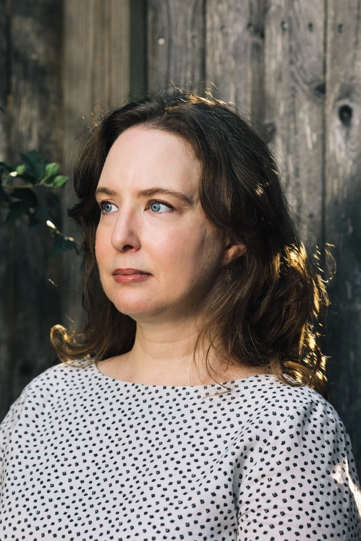A woman with wavy brown hair and blue eyes looking to the side, wearing a white shirt with black polka dots, standing outdoors against a wooden fence.