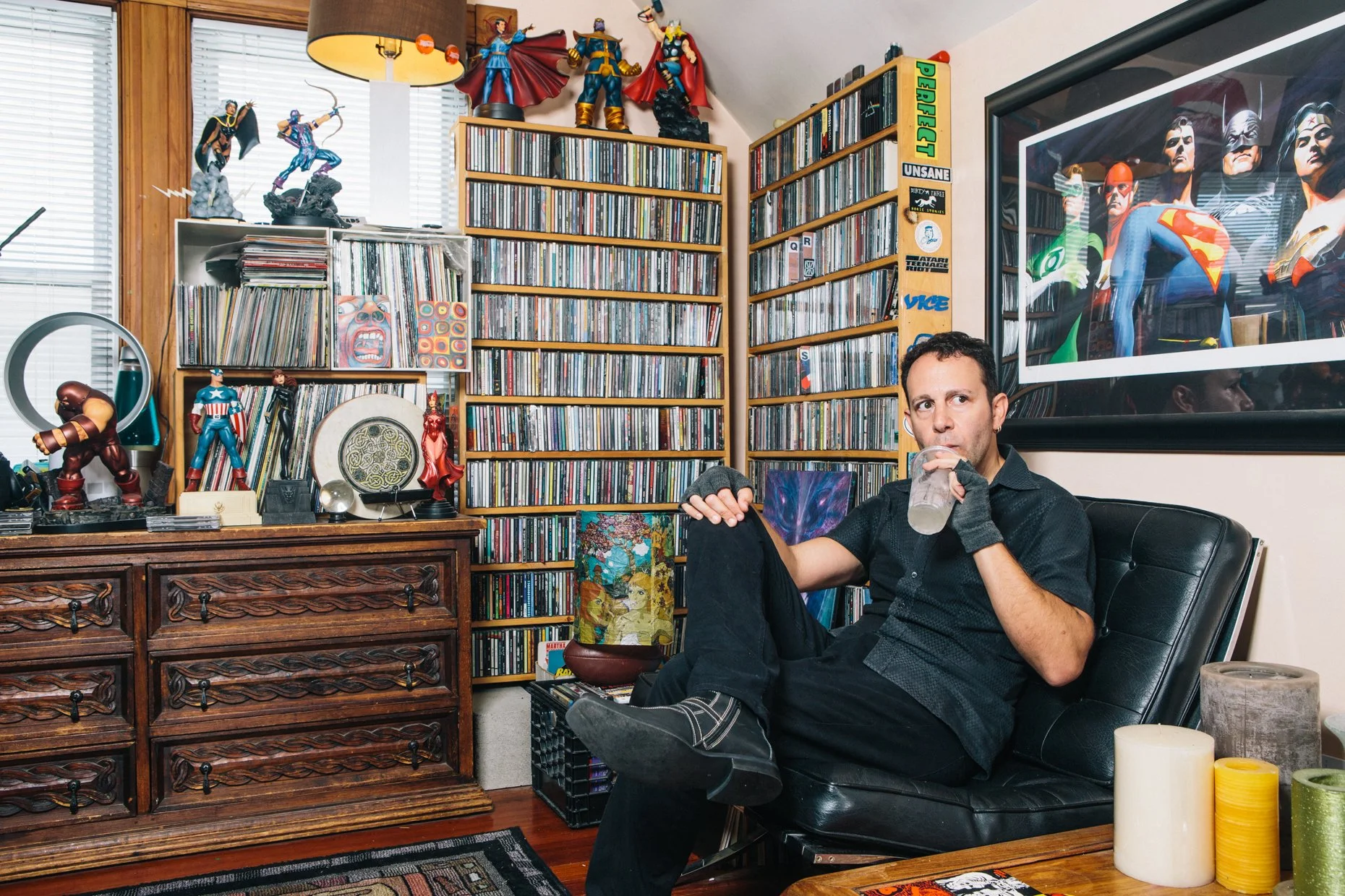 A man sitting in a black leather chair, drinking a beverage from a clear glass, in a room filled with comic book and superhero collectibles, including statues of Marvel characters like Black Panther, Doctor Strange, and Thanos, along with comic books