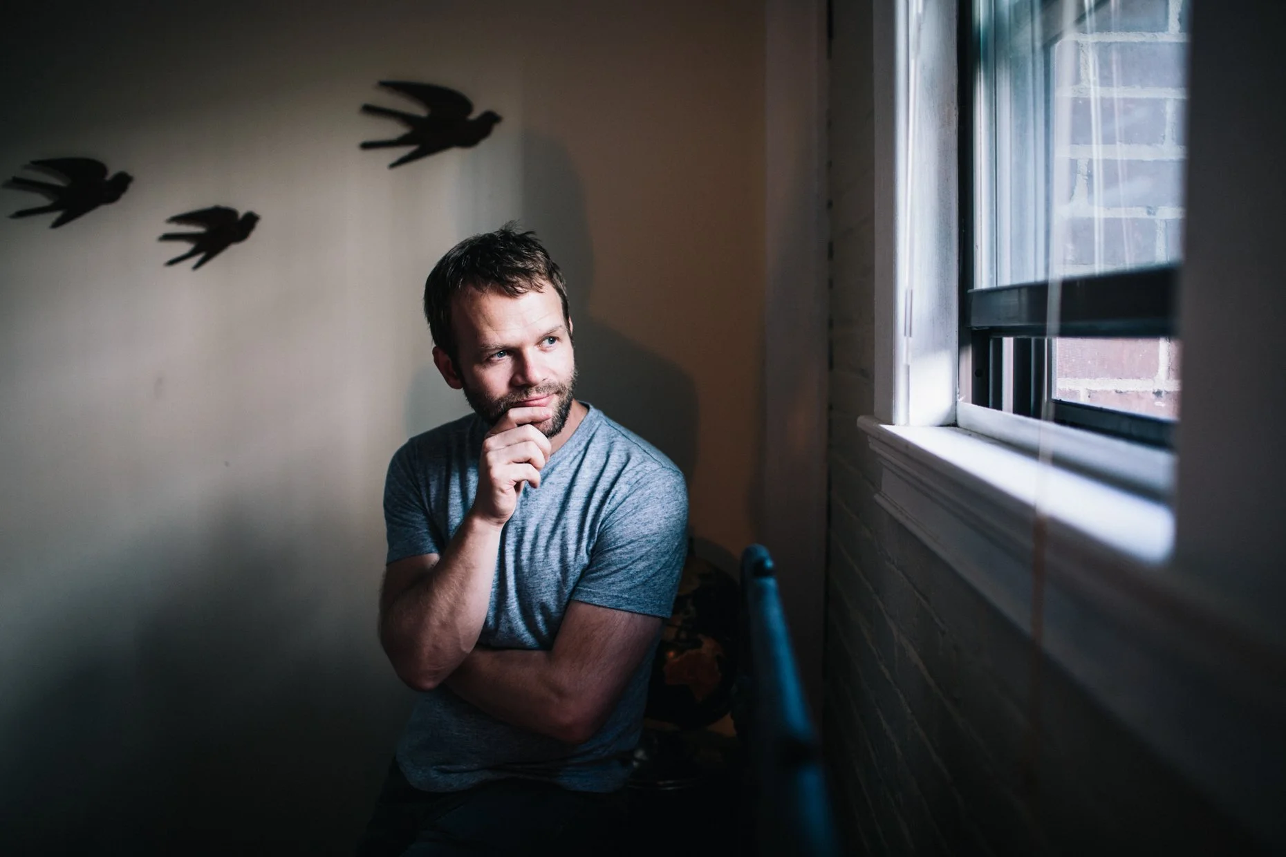 Man sitting by a window with hand on chin, contemplating, with shadow silhouette of flying birds on the wall behind him, natural light coming through window.