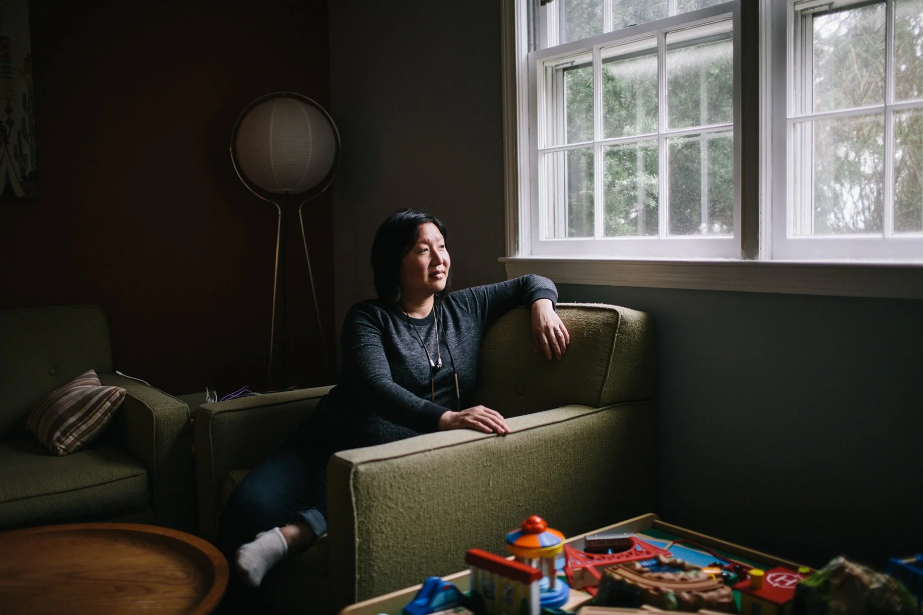 Woman sitting on a green couch near a window, looking outside with a relaxed expression.
