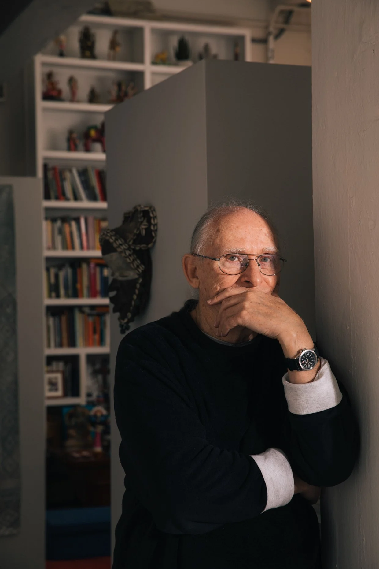 An elderly man with glasses, wearing a black and gray long-sleeve shirt, stands with his hand covering his mouth and nose, leaning against a wall. In the background, there is a white bookshelf filled with books and figurines, and a decorative scarf h