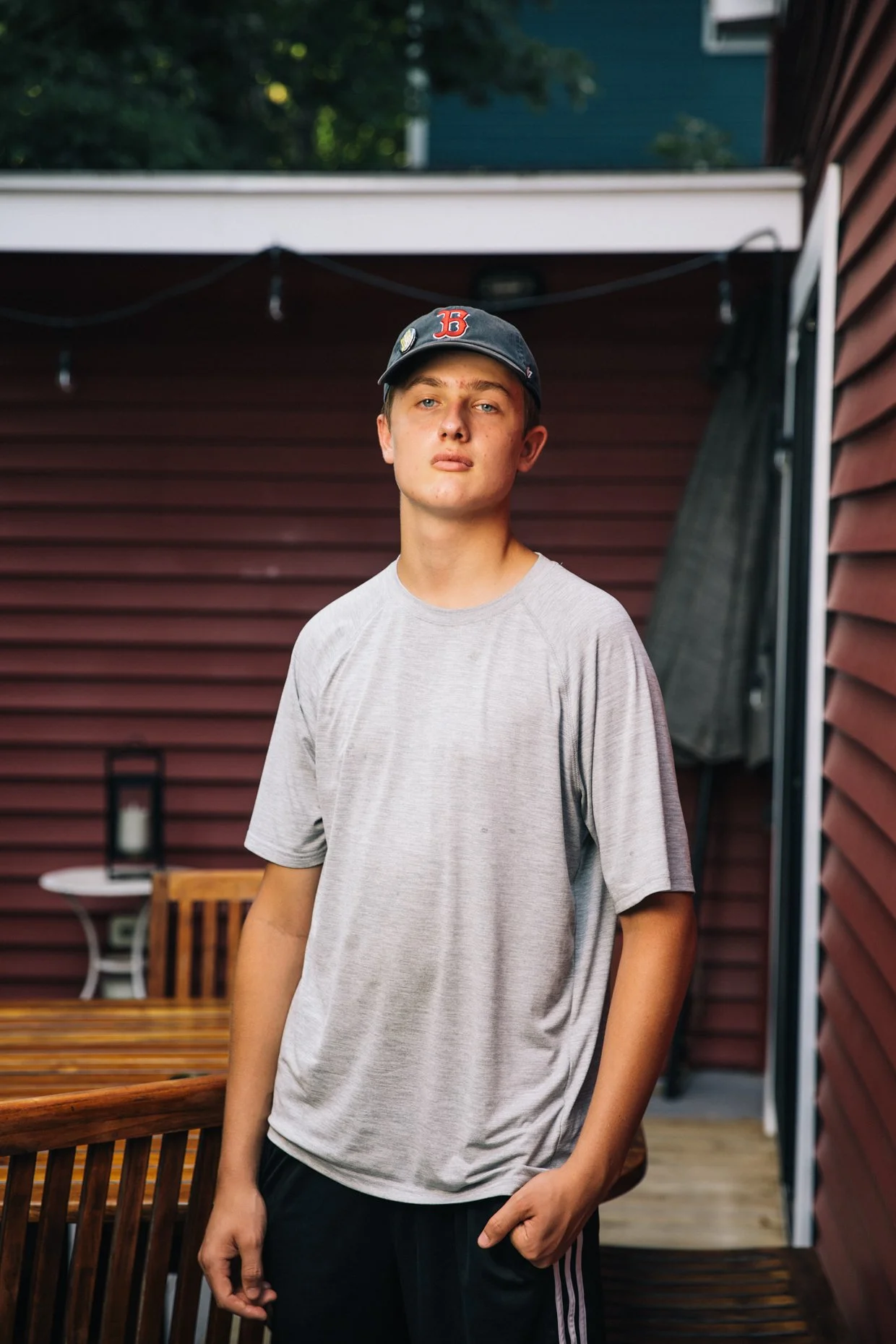 A young man with light skin and blue eyes, wearing a gray t-shirt and black pants with white stripes, stands outdoors in a backyard or patio area with a reddish-brown wooden fence and greenery in the background. He is wearing a dark baseball cap with