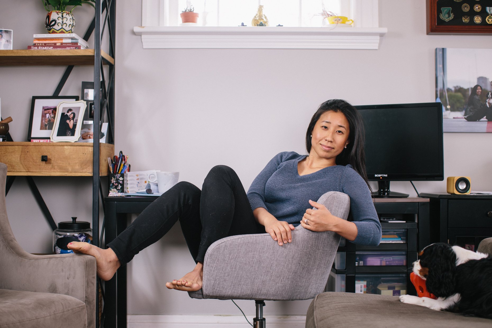 A woman is seated on a gray office chair in a living room, leaning against a beige armchair, with one leg resting on it and the other on the floor. She is smiling and looking at the camera, wearing a blue sweater and black pants. A black and white do