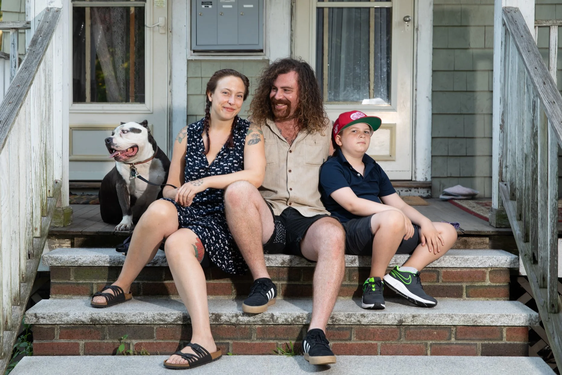 A family of three and their dog sitting on the front porch stairs of a house. The woman has dark hair in braids and is wearing a black and white dress. The man has long curly hair and a beard, wearing a beige shirt and black shorts. The boy is wearin