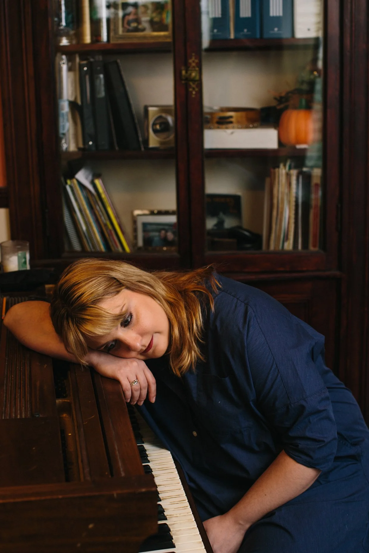 A woman with reddish-blonde hair resting her head on her arm on a piano, looking thoughtful.