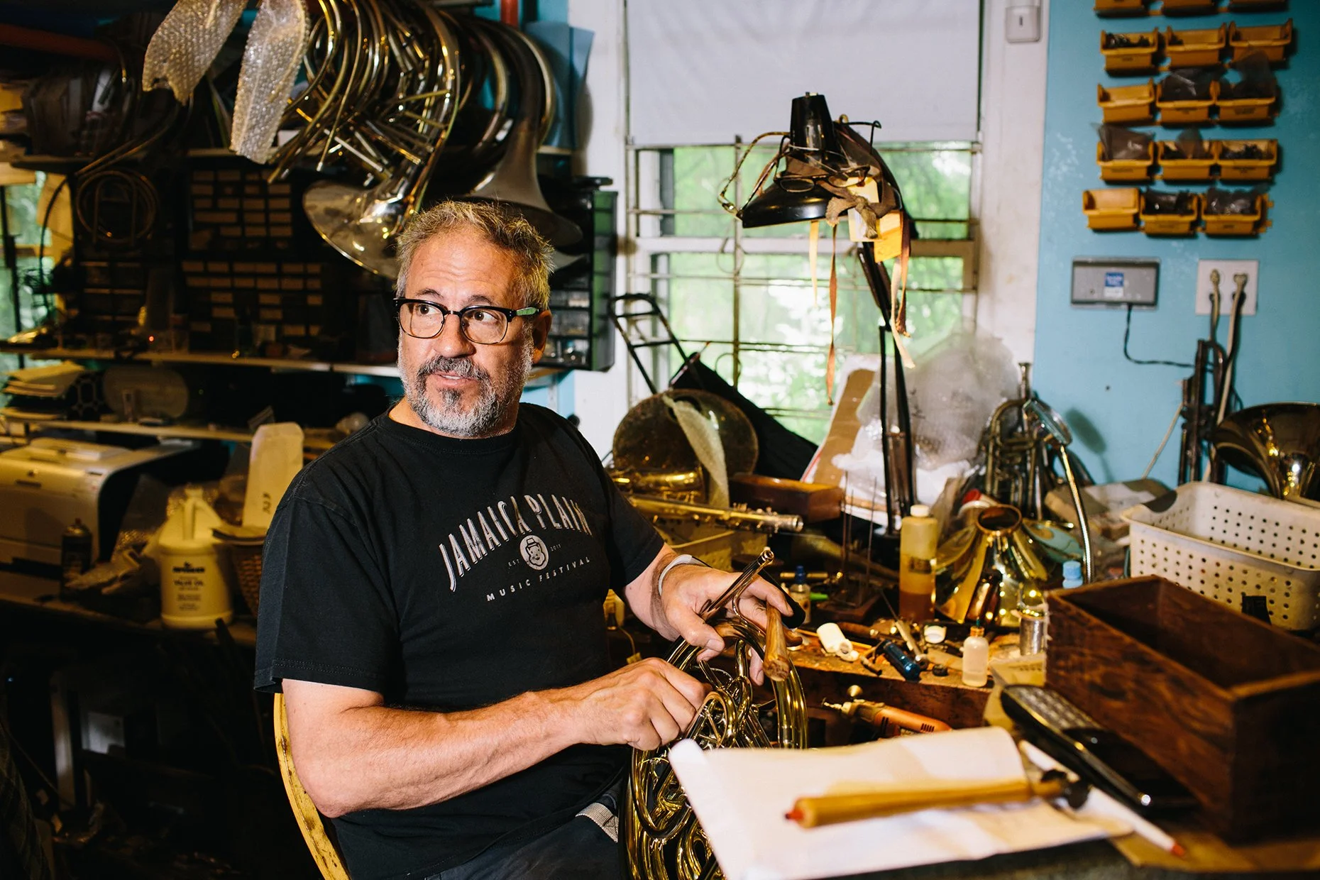 A man with glasses and a beard working with a French horn in a cluttered workshop filled with musical instruments and tools.