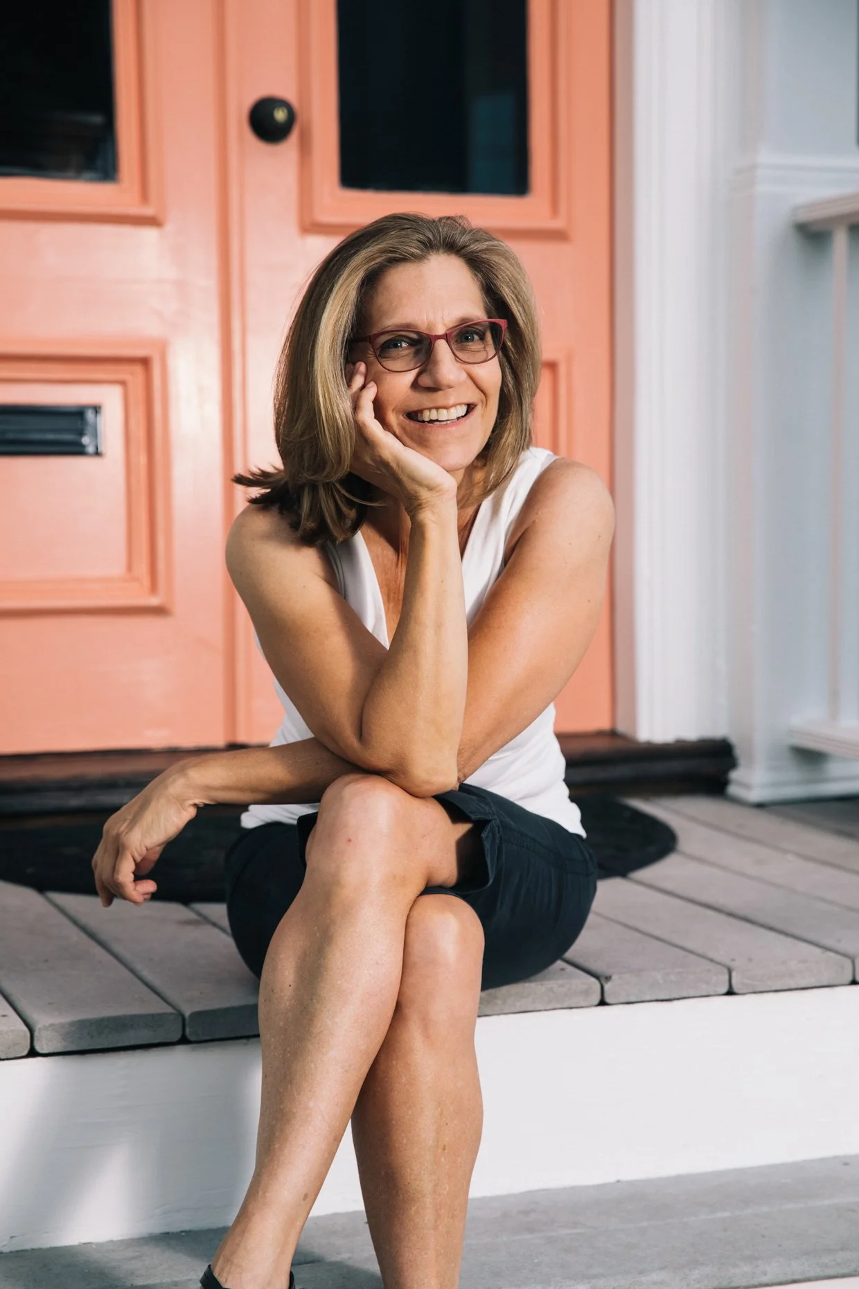 A woman with shoulder-length hair, wearing glasses, a white sleeveless top, and black shorts, is sitting on the porch of a house with a pink door, smiling and resting her head on her hand.