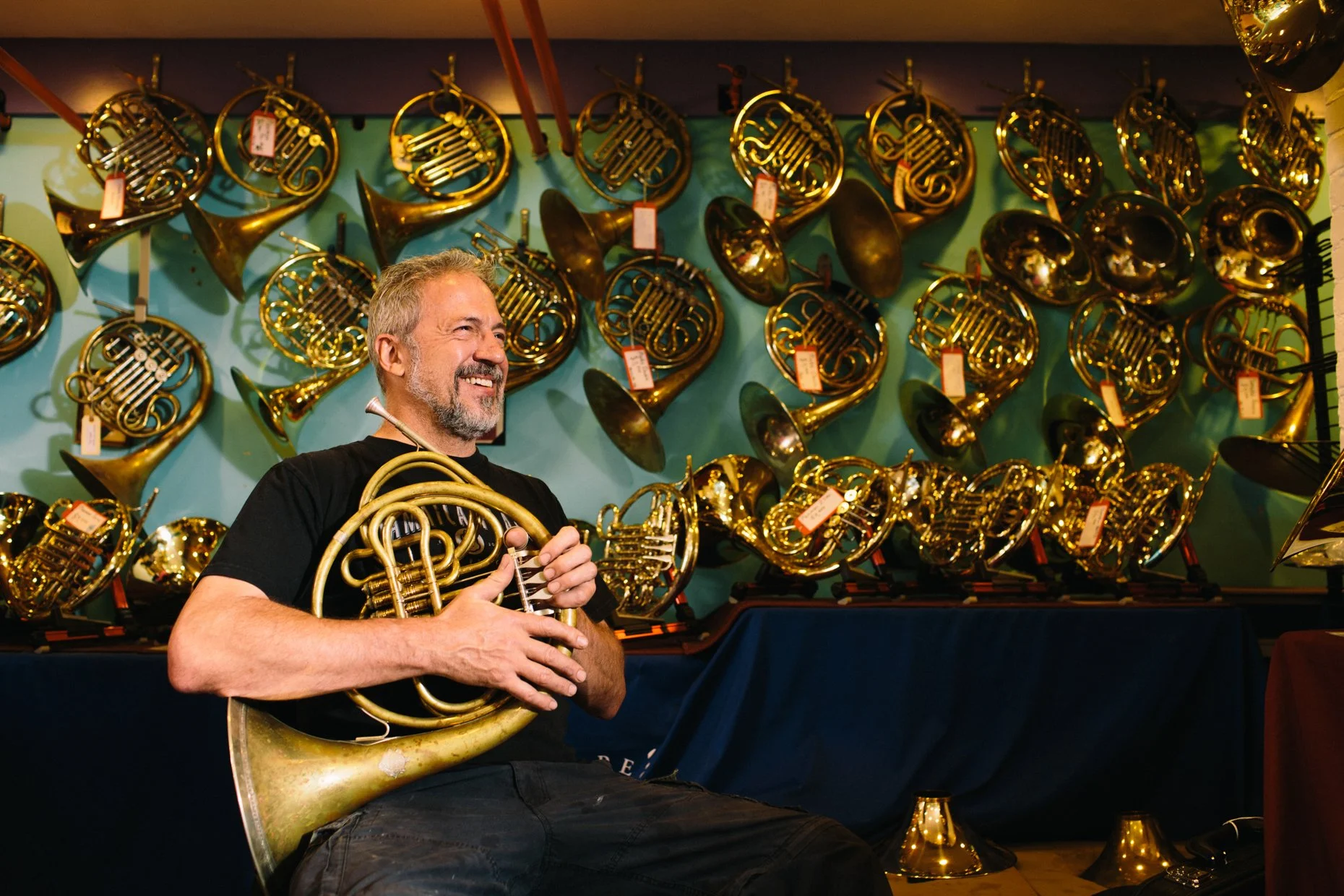 A man with gray hair and a beard holding a French horn, standing in front of a wall display of various brass musical instruments, mostly French horns, in a music shop or store.