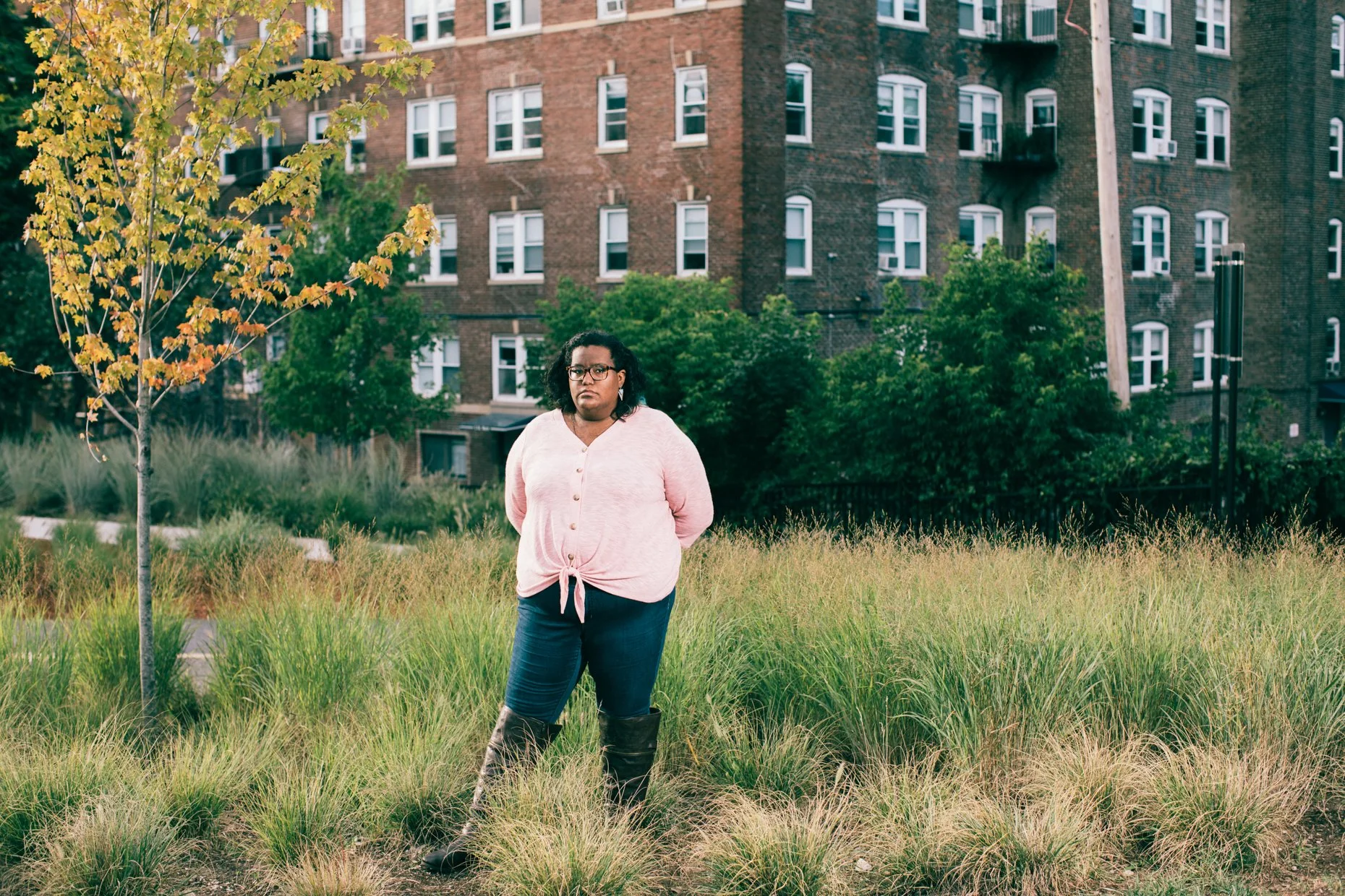 A woman standing in a grassy urban park with trees and a brick apartment building in the background. She is wearing glasses, a light pink sweater, dark pants, and black boots. Her hands are behind her back and she is looking at the camera.