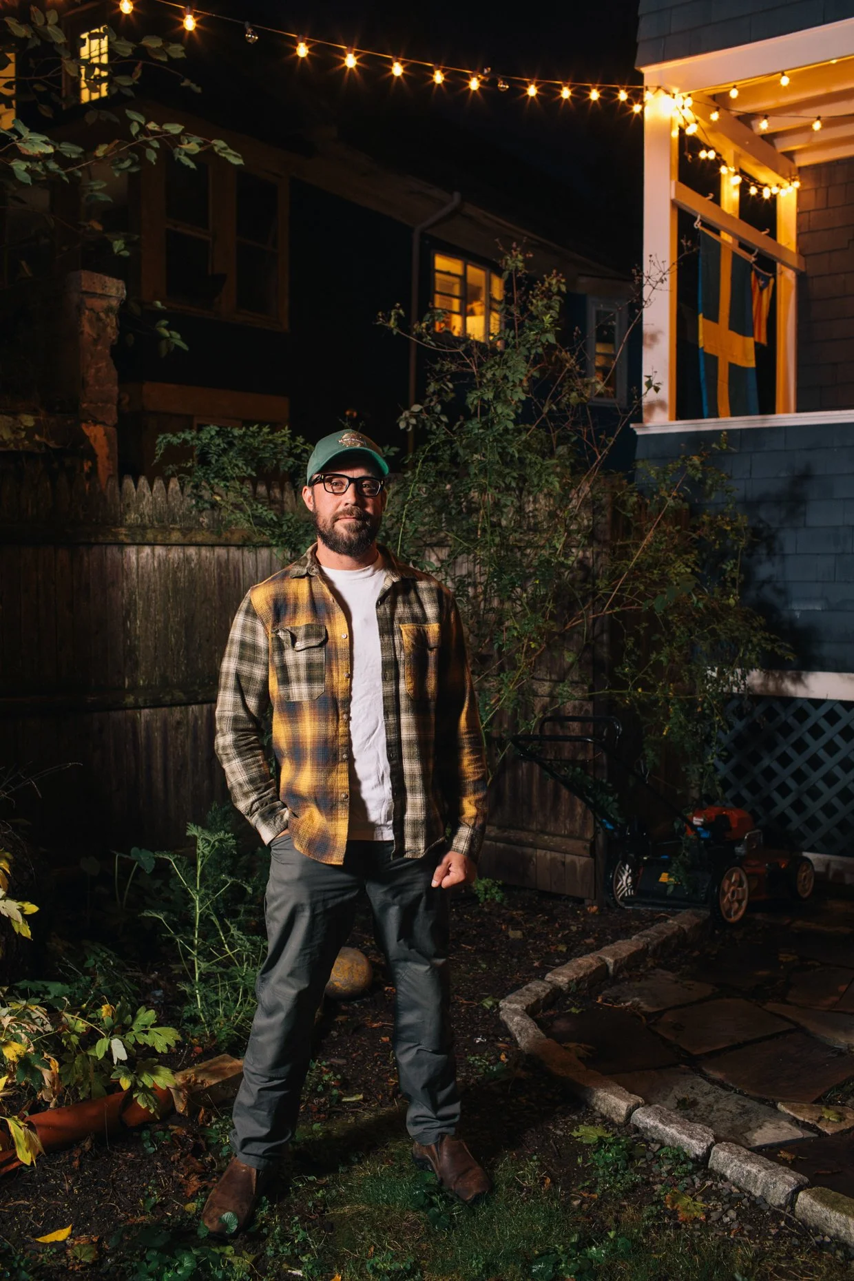 A man with glasses, a beard, wearing a green cap, a plaid shirt, and gray pants, standing in a backyard at night with string lights overhead, plants, a wooden fence, and a house with Swedish flags hanging in the background.