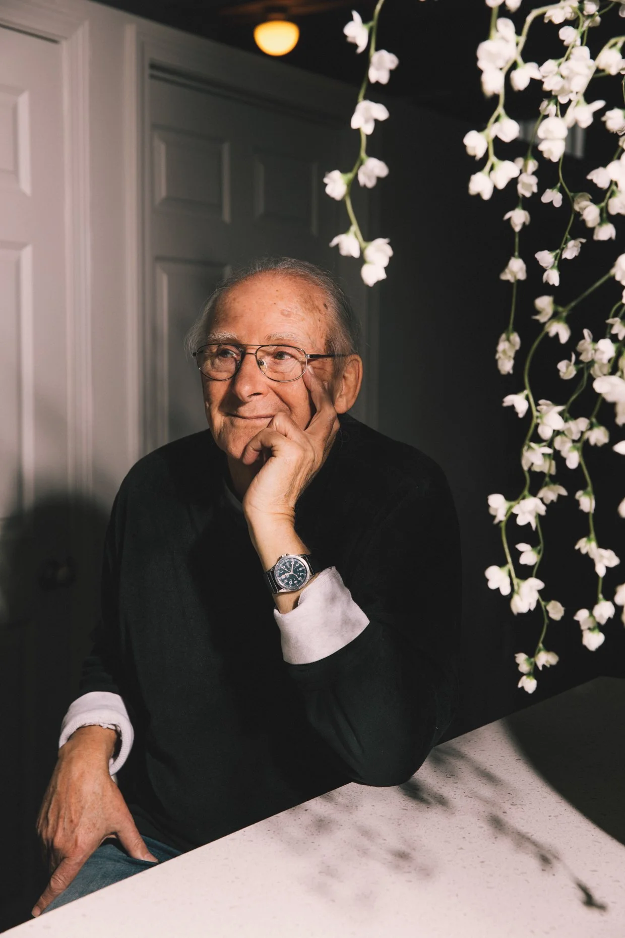 An elderly man wearing glasses, a black sweater, and a wristwatch sitting at a table with a thoughtful expression, resting his chin on his hand, near white flowers hanging from the ceiling.