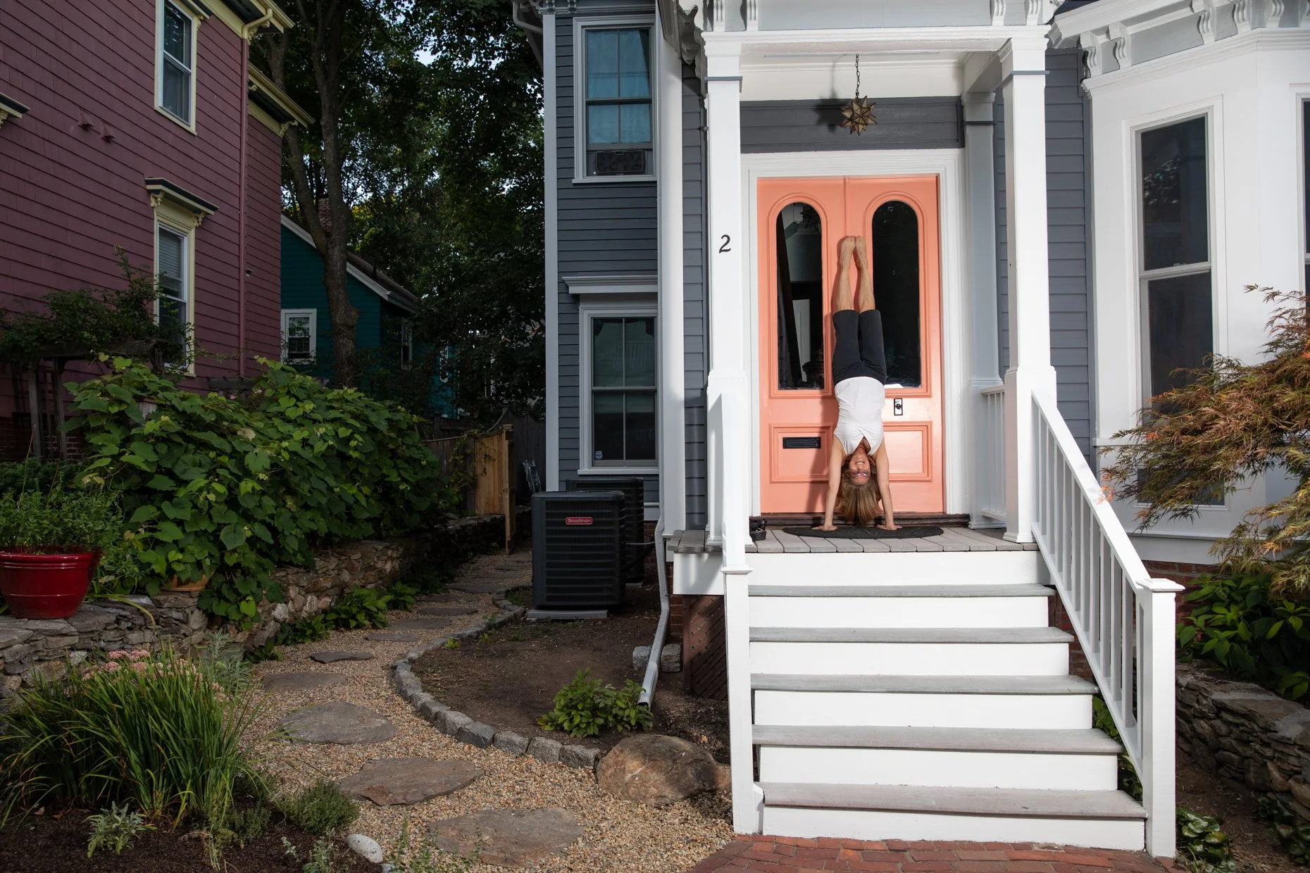 A woman doing a handstand on the porch of a blue house with a coral front door, surrounded by a garden with rocks, plants, and a pathway.