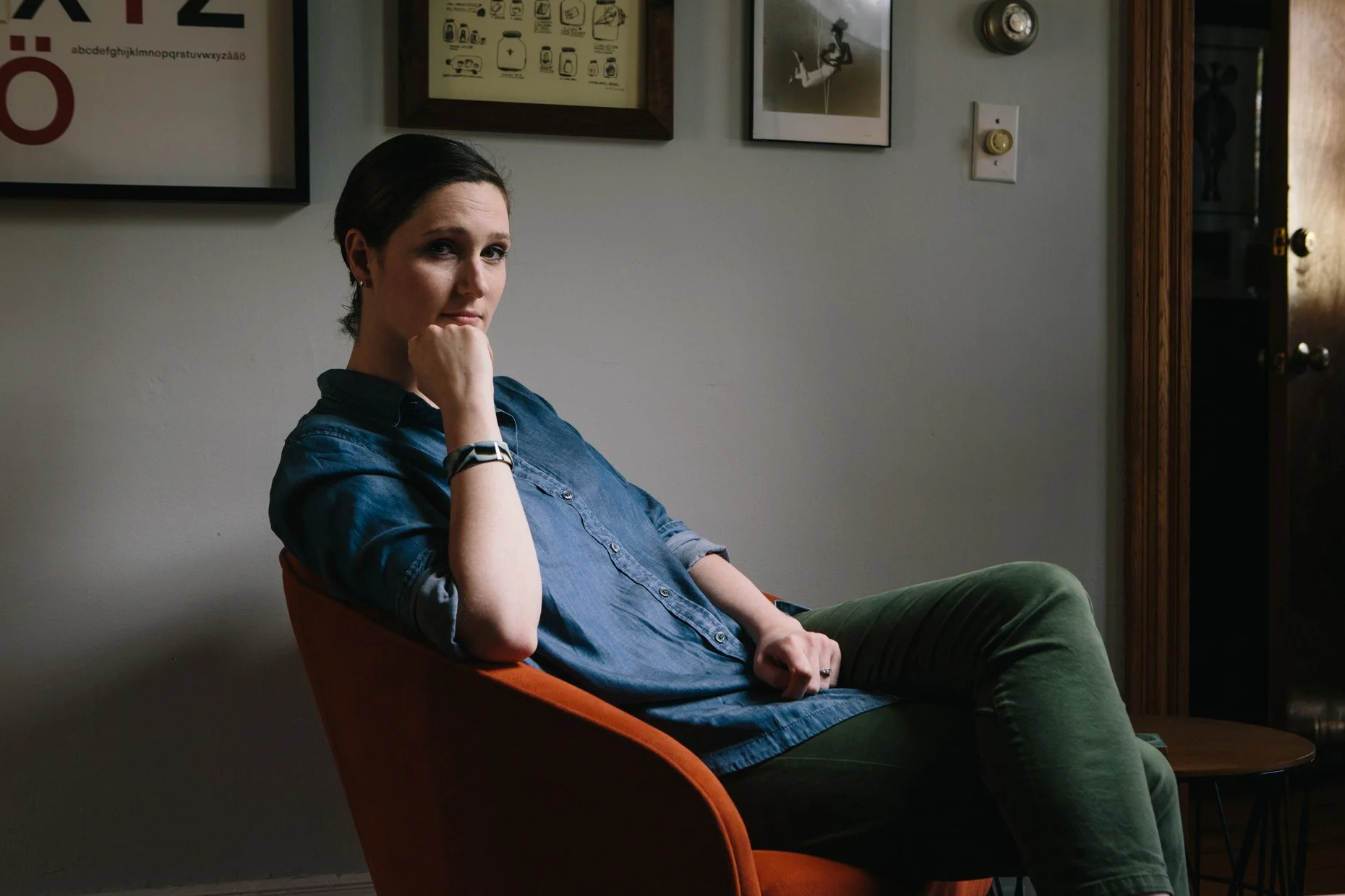 A woman sitting in an orange chair with a contemplative expression, wearing a denim shirt and green pants, in a room with framed pictures and artwork on the wall.