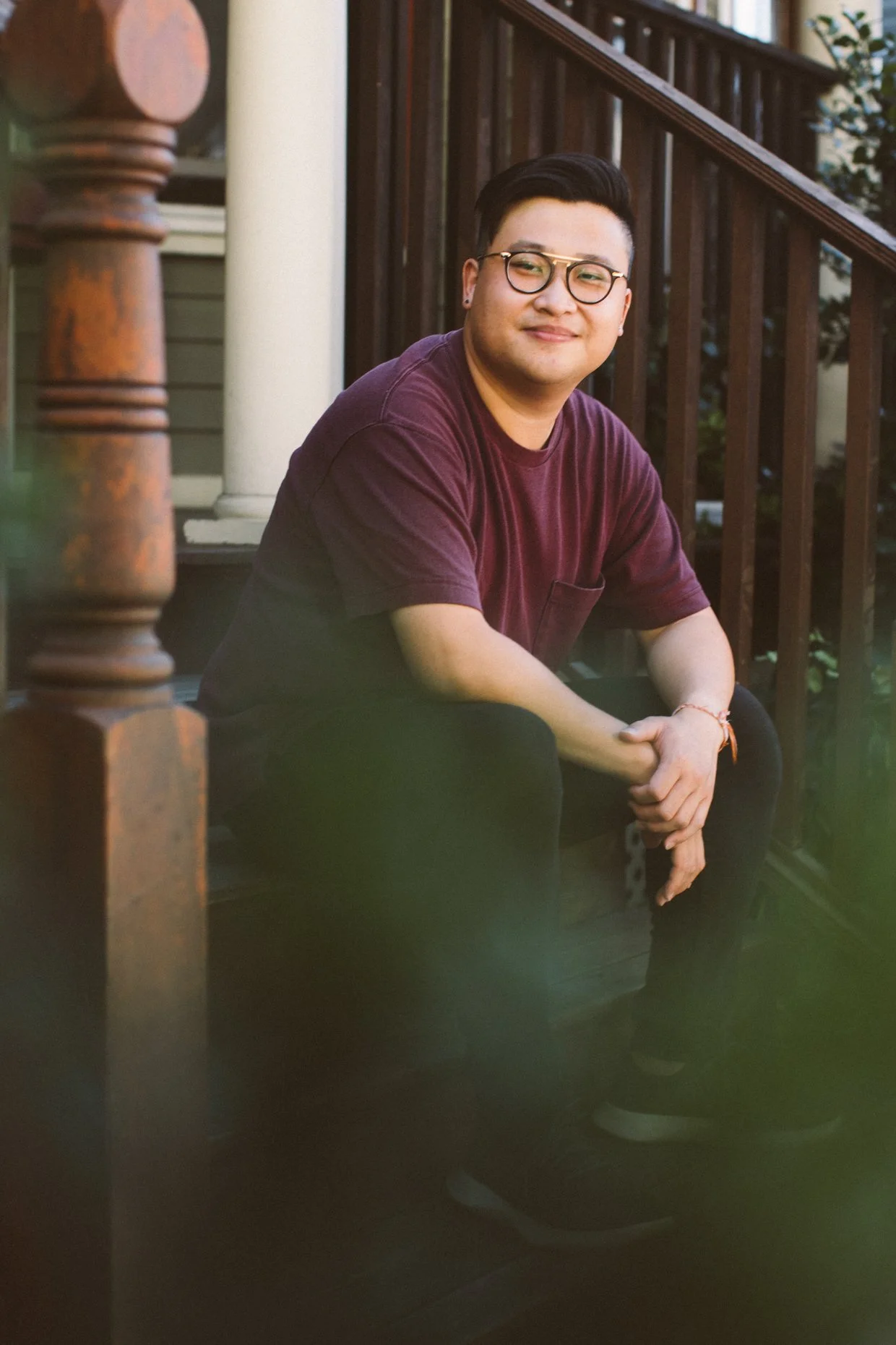 A young man with glasses and earrings sitting on a porch with wooden railings, smiling at the camera, with a house and greenery in the background.