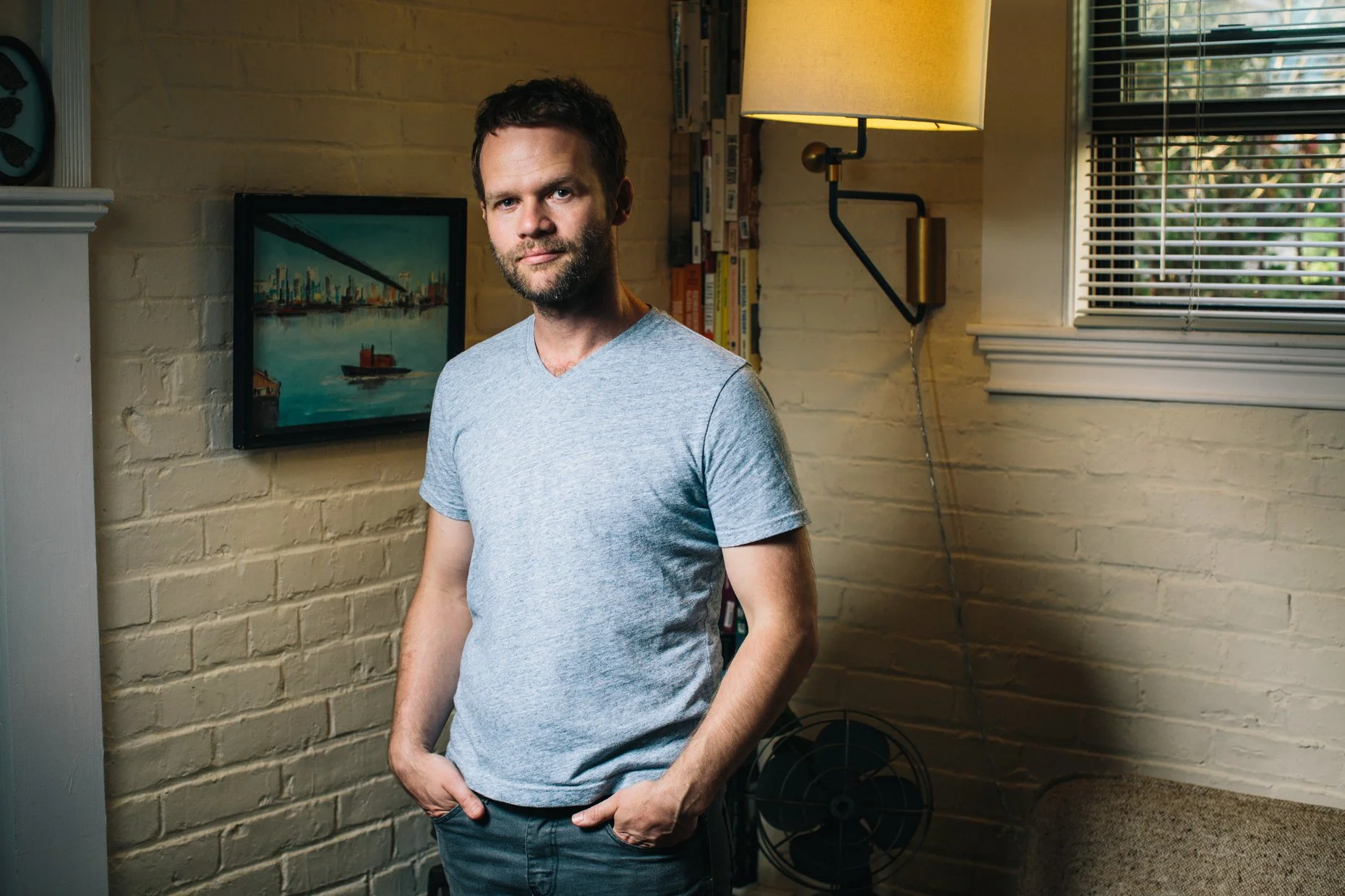 A man standing indoors near a brick wall, with a painting of a city skyline and a boat on water behind him, and a window with blinds to his right. He's wearing a light gray t-shirt, has a beard, and is looking at the camera.