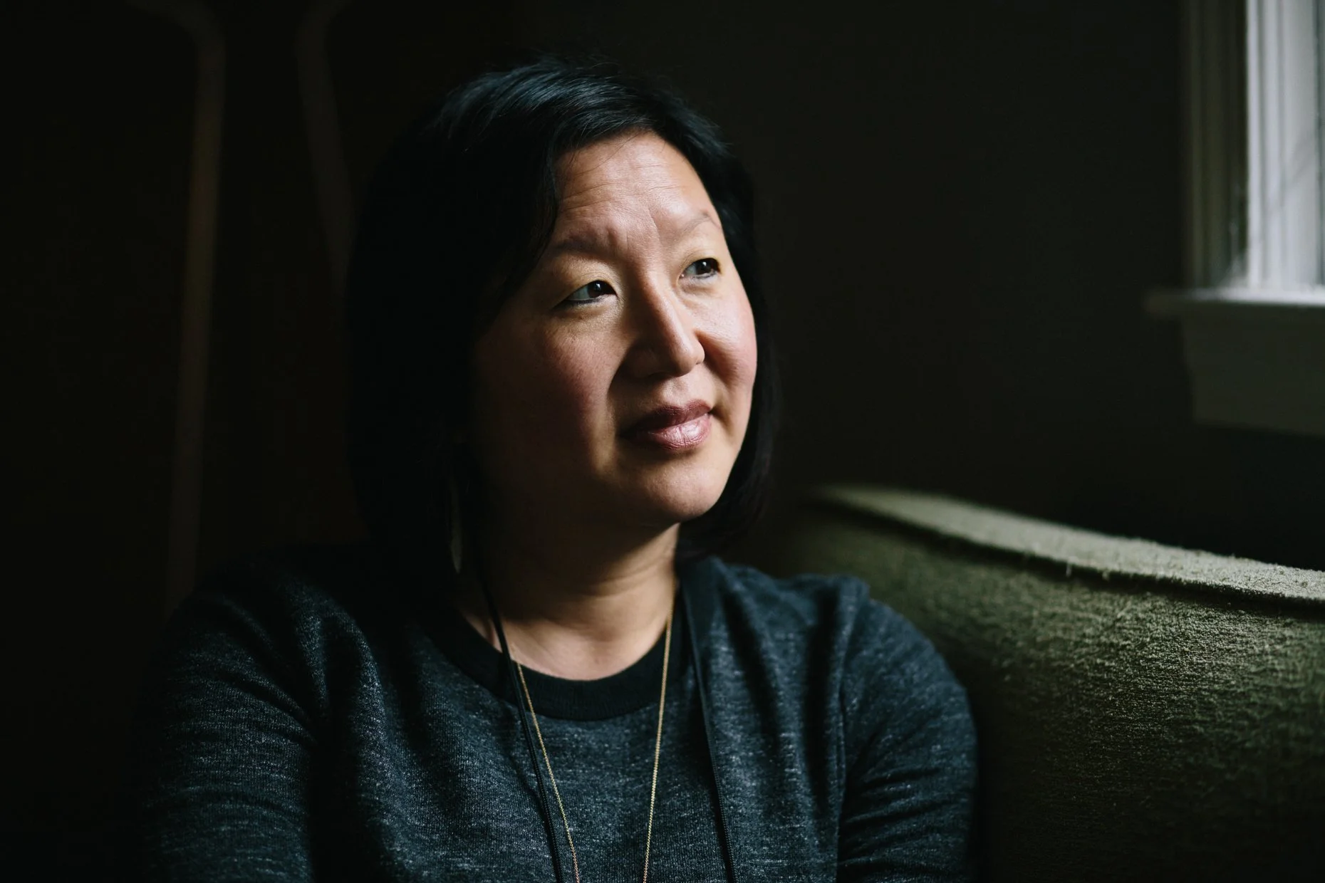 A woman with short black hair sitting on a couch near a window, looking thoughtfully.