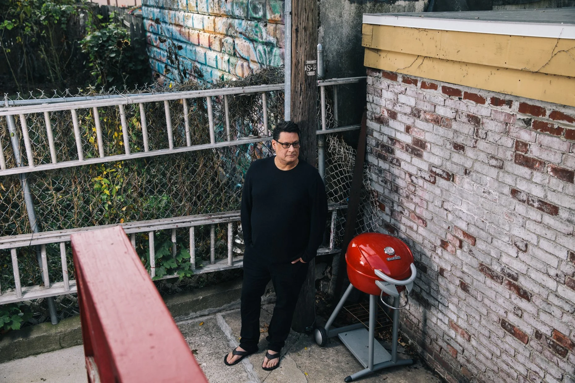A man standing outdoors next to a brick wall with a red barbecue grill nearby. He is wearing glasses, a black shirt, and flip-flops, and is looking slightly to the right with a serious expression. Behind him, there is a chain-link fence with overgrow