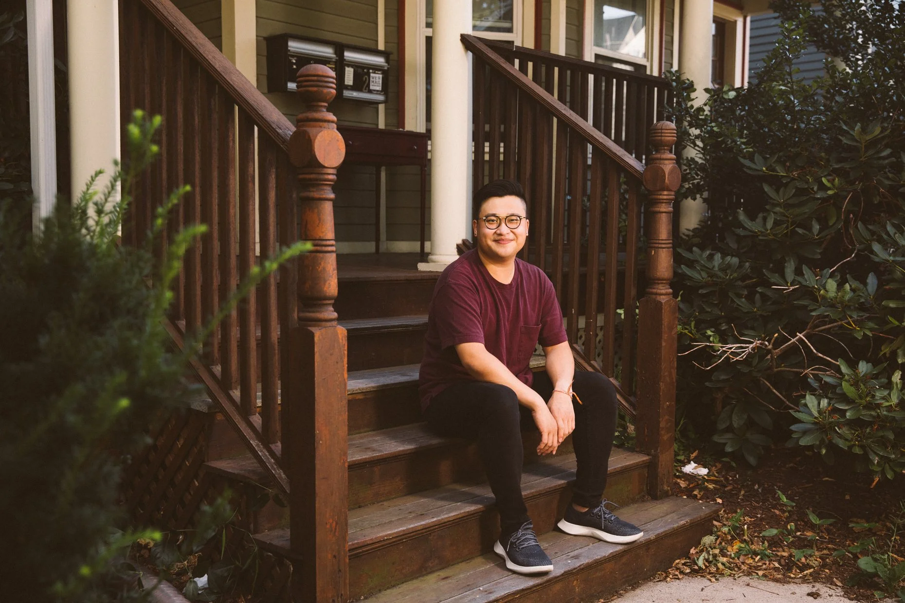 A young man with glasses, wearing a maroon shirt and black pants, sitting on wooden stairs outside a house, surrounded by greenery.