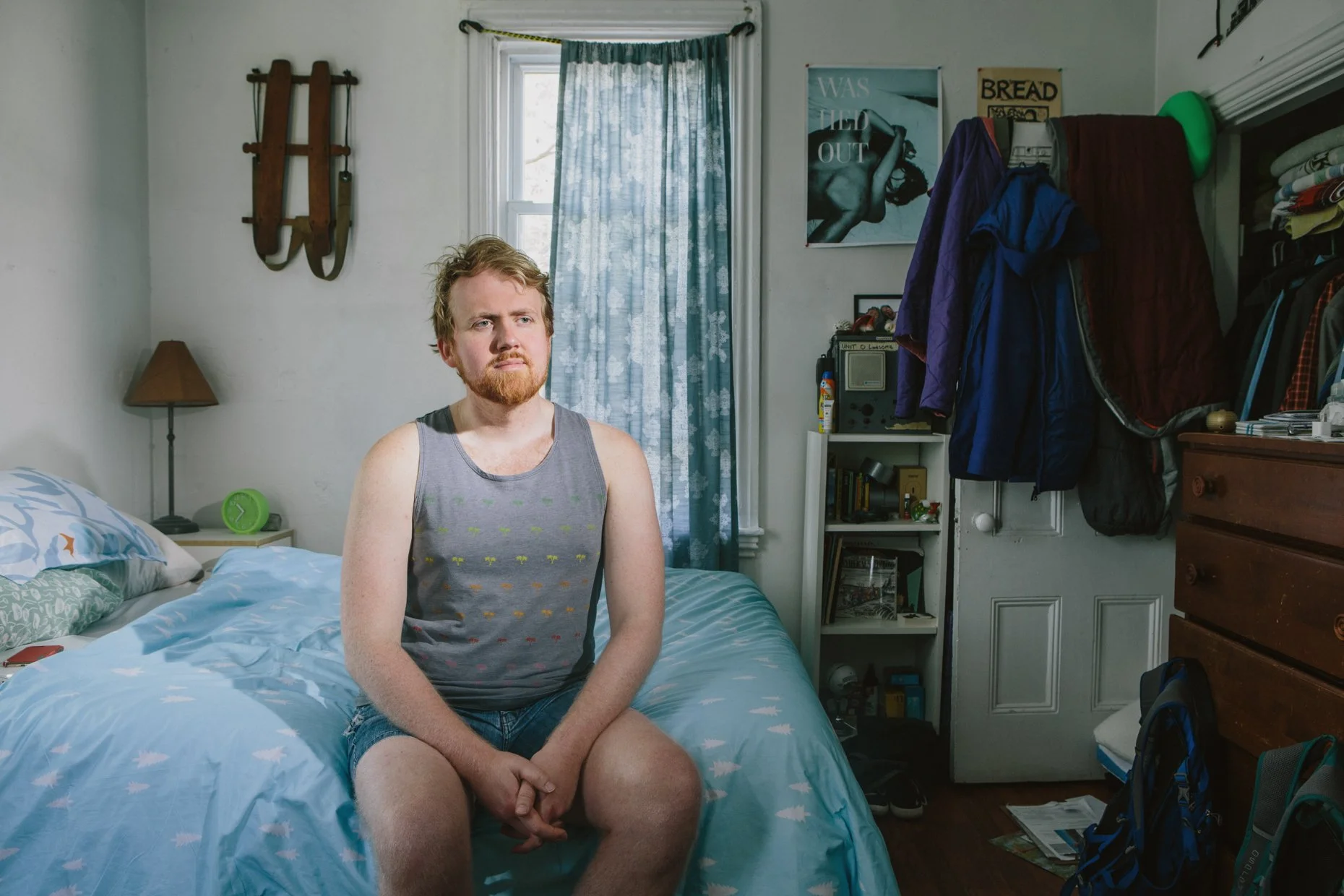 A young man with red hair and beard sits on a bed in a messy bedroom, wearing a gray tank top and shorts, looking contemplative. The room has a window with blue curtains, a small nightstand with a green alarm clock, a lamp, and a wall decor that incl