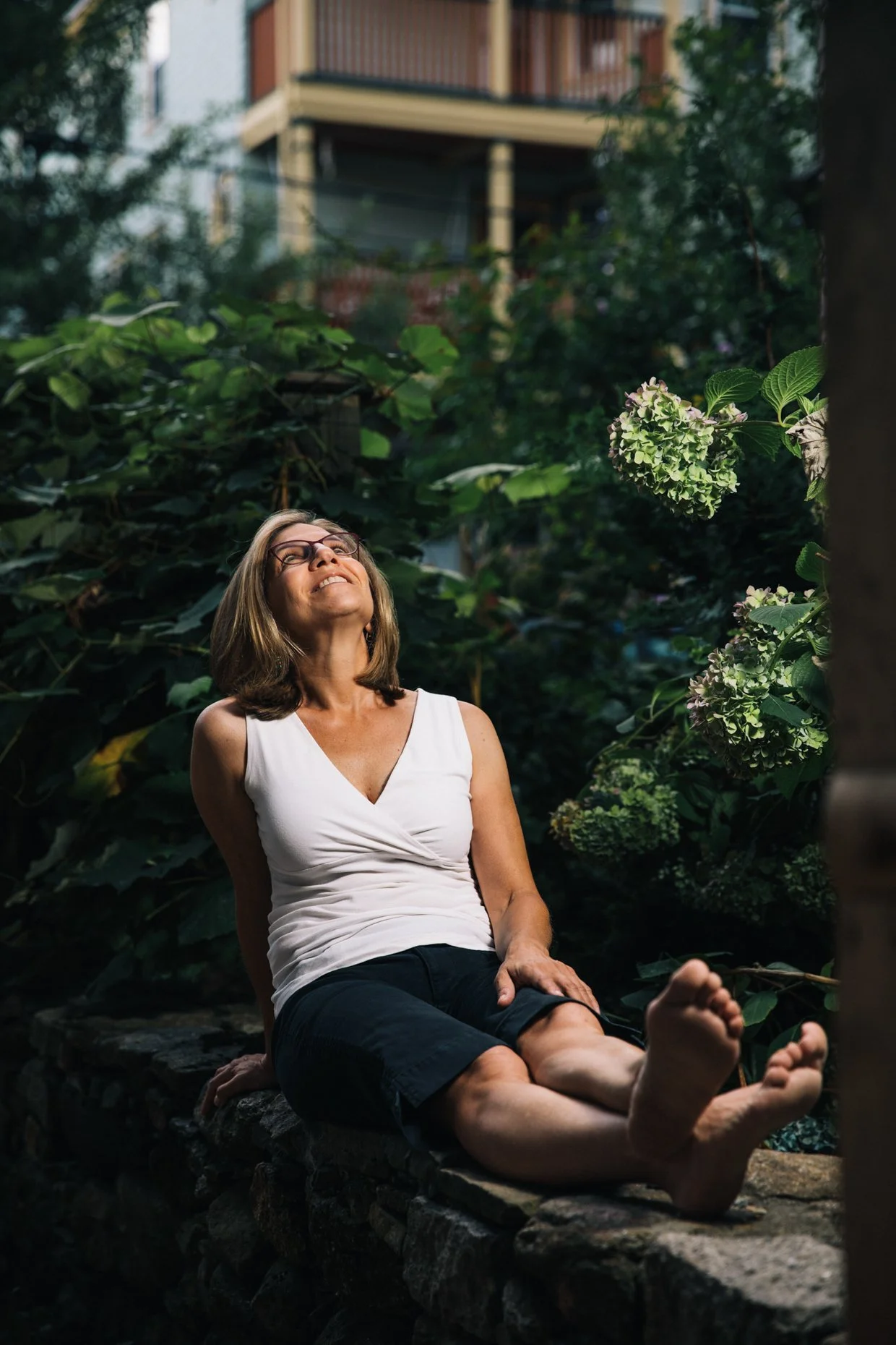 A woman with glasses and shoulder-length hair is sitting on a stone ledge in a garden with lush green plants and hydrangea flowers, smiling and looking upwards.