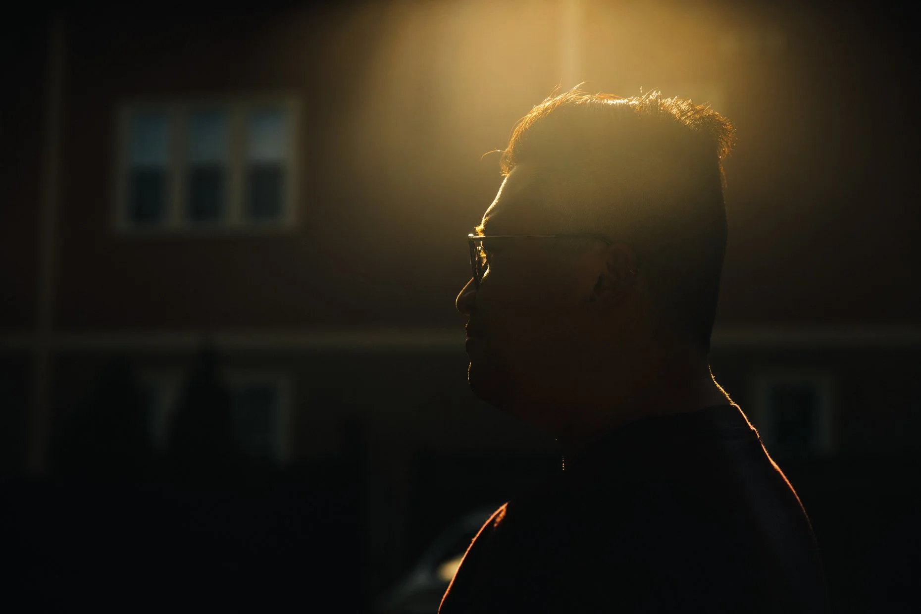 Profile of a person with short hair and glasses, illuminated by warm light from behind, with a dark blurred background.