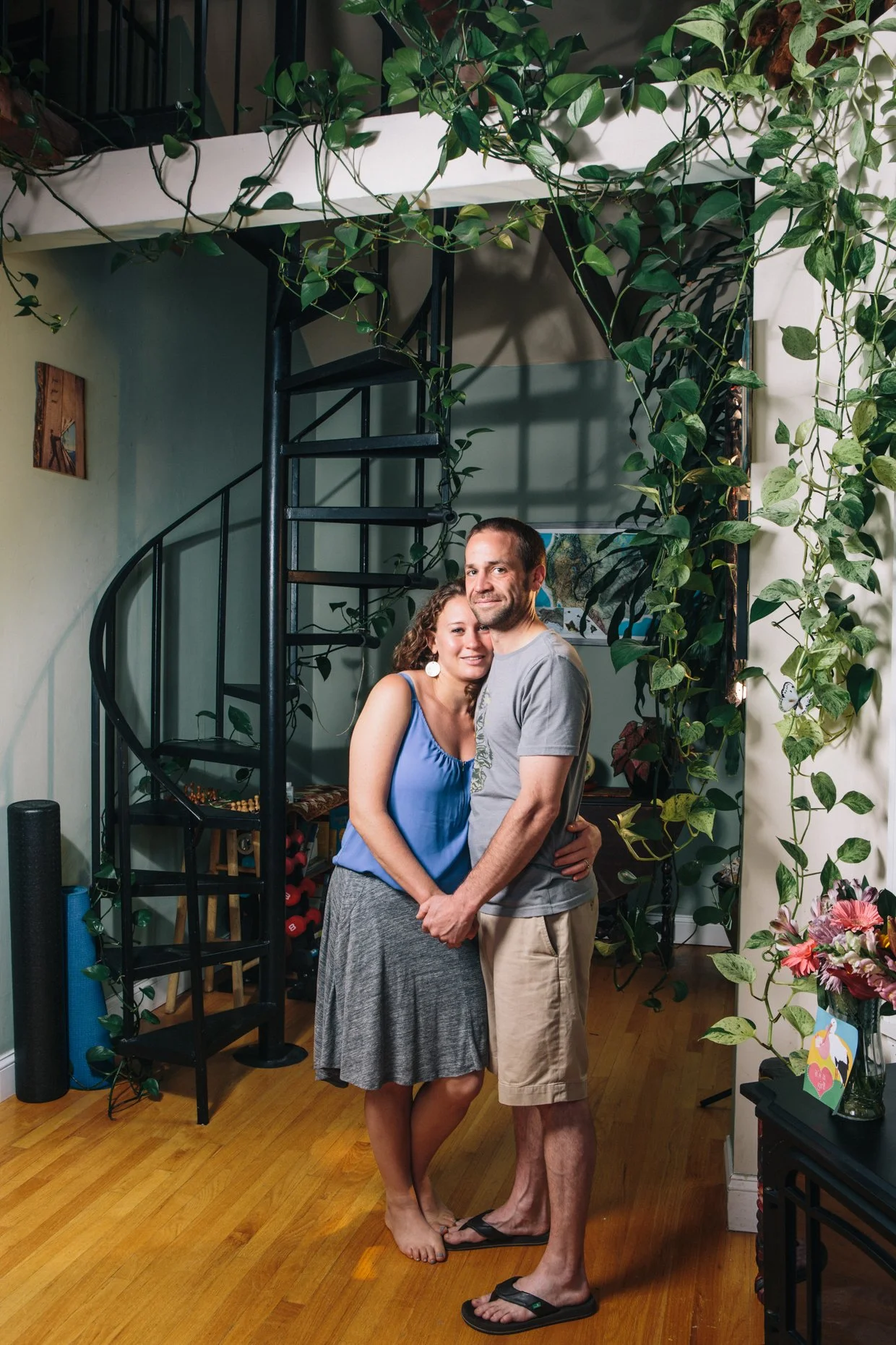 A couple stands close together inside a home, embracing each other and smiling at the camera. They are barefoot and dressed casually. The background includes a spiral staircase, lush green plants, and a corner with various decorations and pictures on