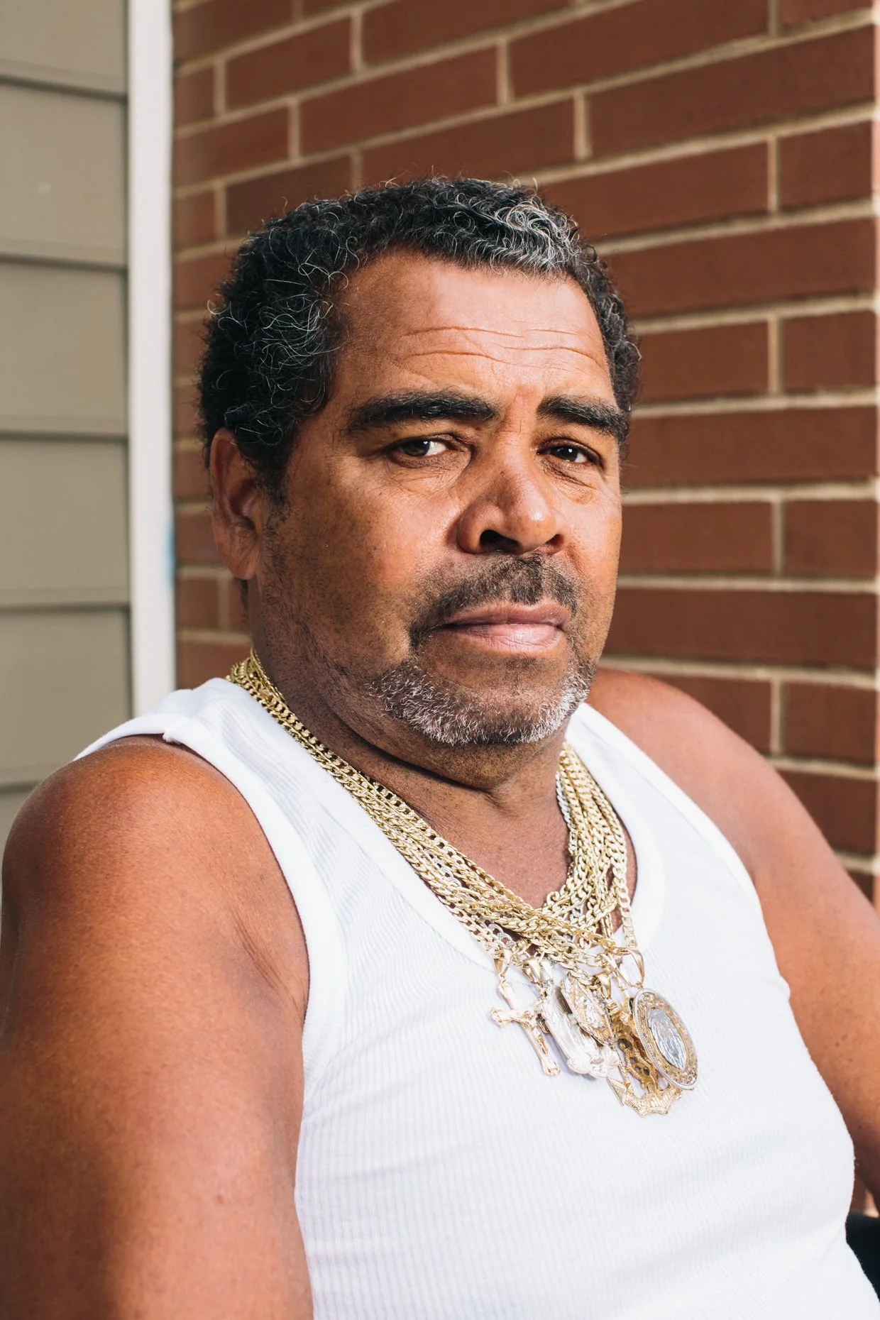 A middle-aged man with dark, curly hair and a beard, wearing a white sleeveless shirt and layered gold chains around his neck, sitting outside against a brick wall.