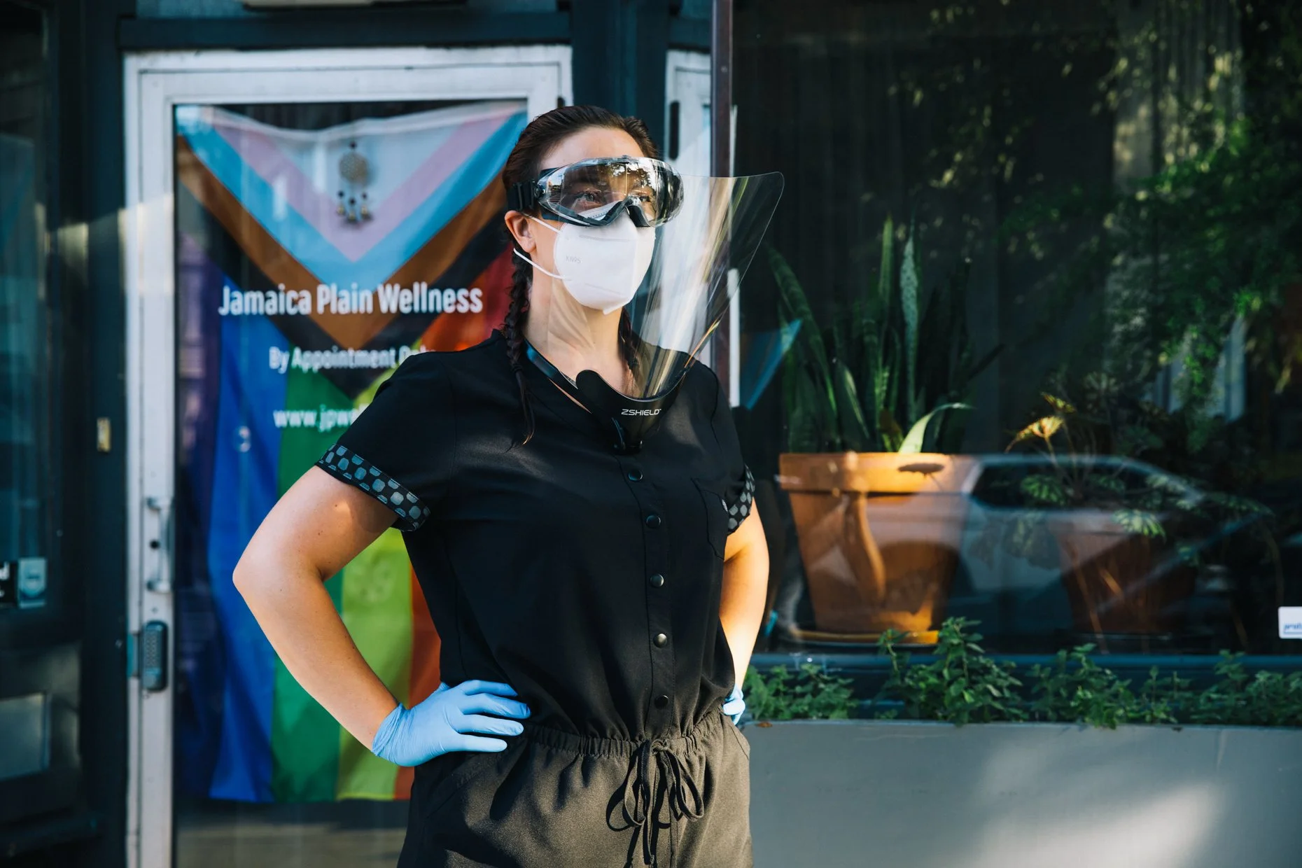 A woman wearing protective gear, including a mask, goggles, and gloves, standing outdoors in front of a storefront with a sign that reads 'Jamaica Plain Wellness'. She has braided hair and is dressed in a black shirt and gray pants with a drawstring.