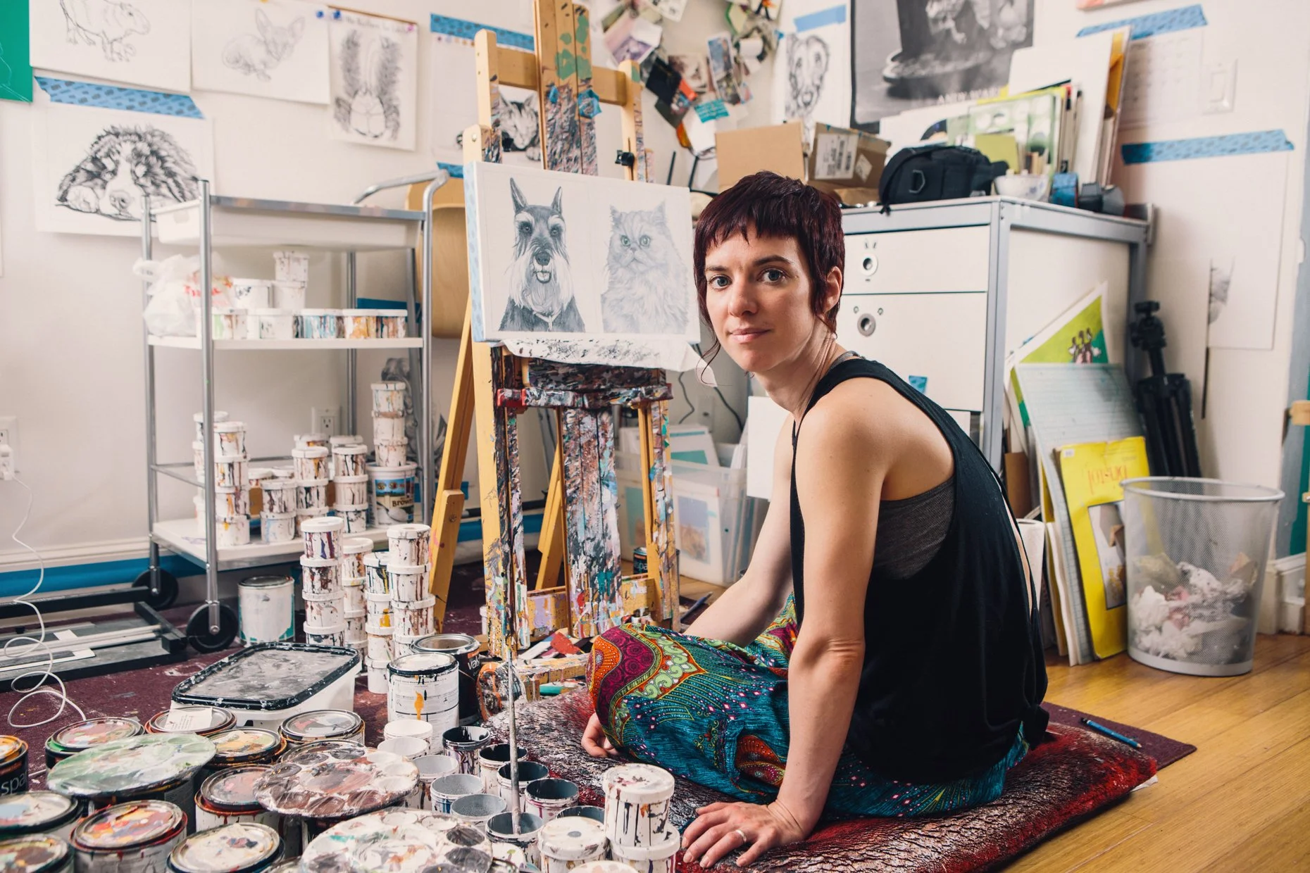 A woman with short dark hair sitting on a rug in art studio, surrounded by paint cans, brushes, and artwork, with a canvas of a dog and cat behind her.