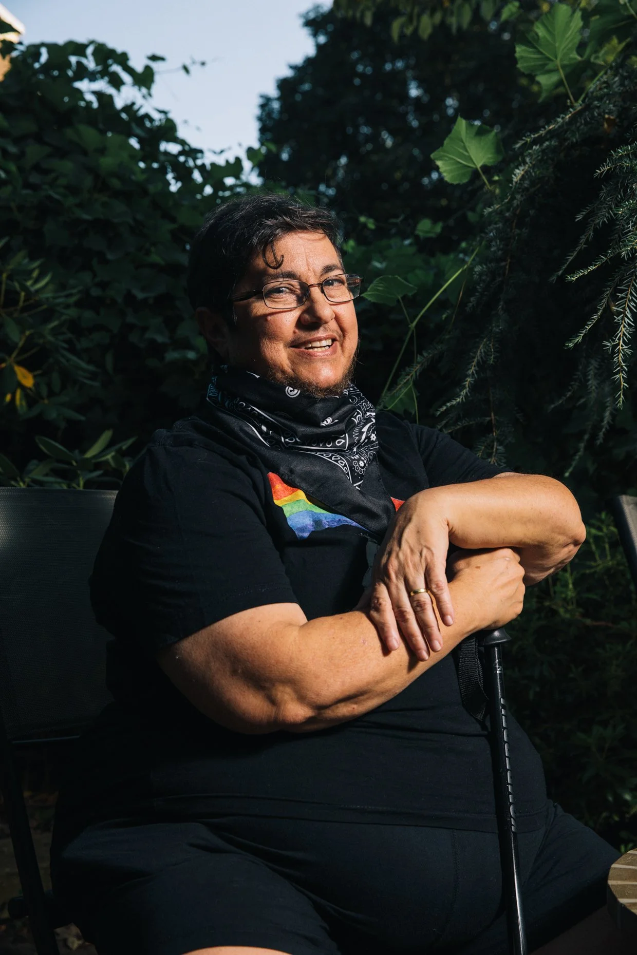 A person with short dark hair, glasses, and a rainbow pride T-shirt sitting outdoors among green foliage, smiling at the camera with arms crossed and holding a cane.