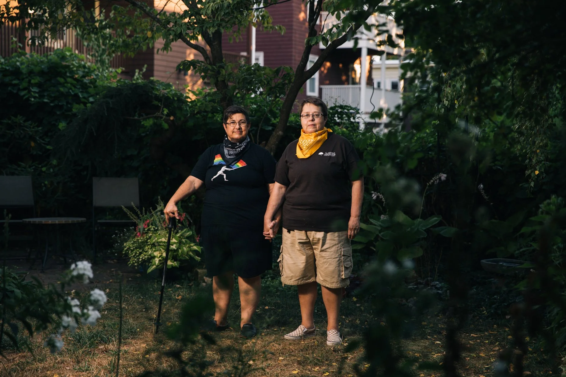 Two people, one with a cane and rainbow pride flag on their shirt and the other with a yellow bandana, standing together in a garden surrounded by greenery.