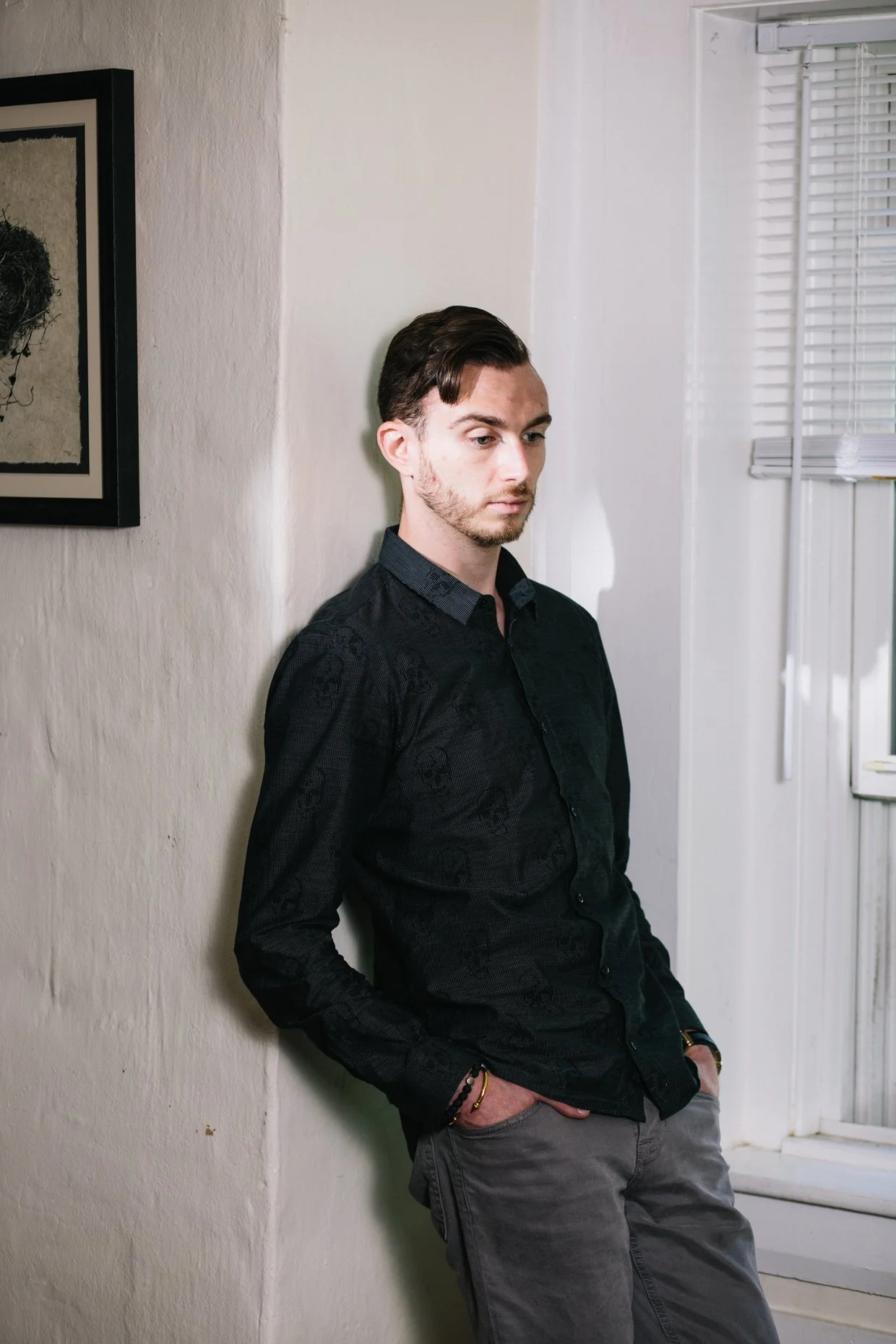 A young man with dark hair and a beard is leaning against a white wall near a window with blinds, wearing a black patterned shirt and gray pants, looking down with a pensive expression.