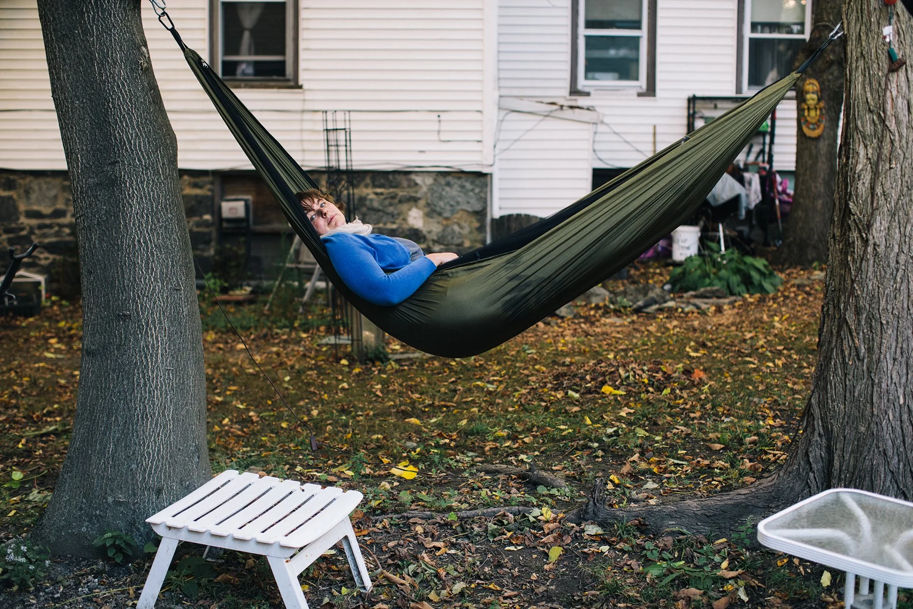 Person lying in a hammock between two trees in a backyard with fallen leaves, a white bench in the foreground, and a house with white siding in the background.