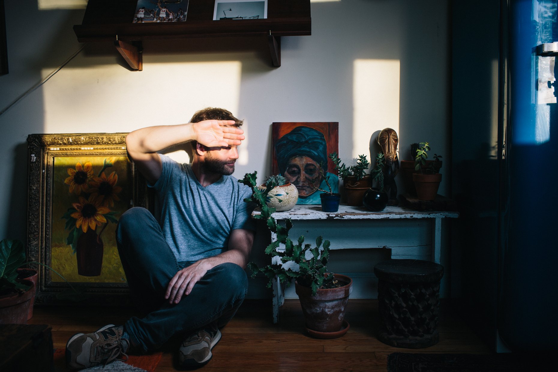 A man with a beard and casual clothing sitting cross-legged on the floor in a room with various plants, artwork, and decorative items, shielding his eyes from sunlight with his hand.
