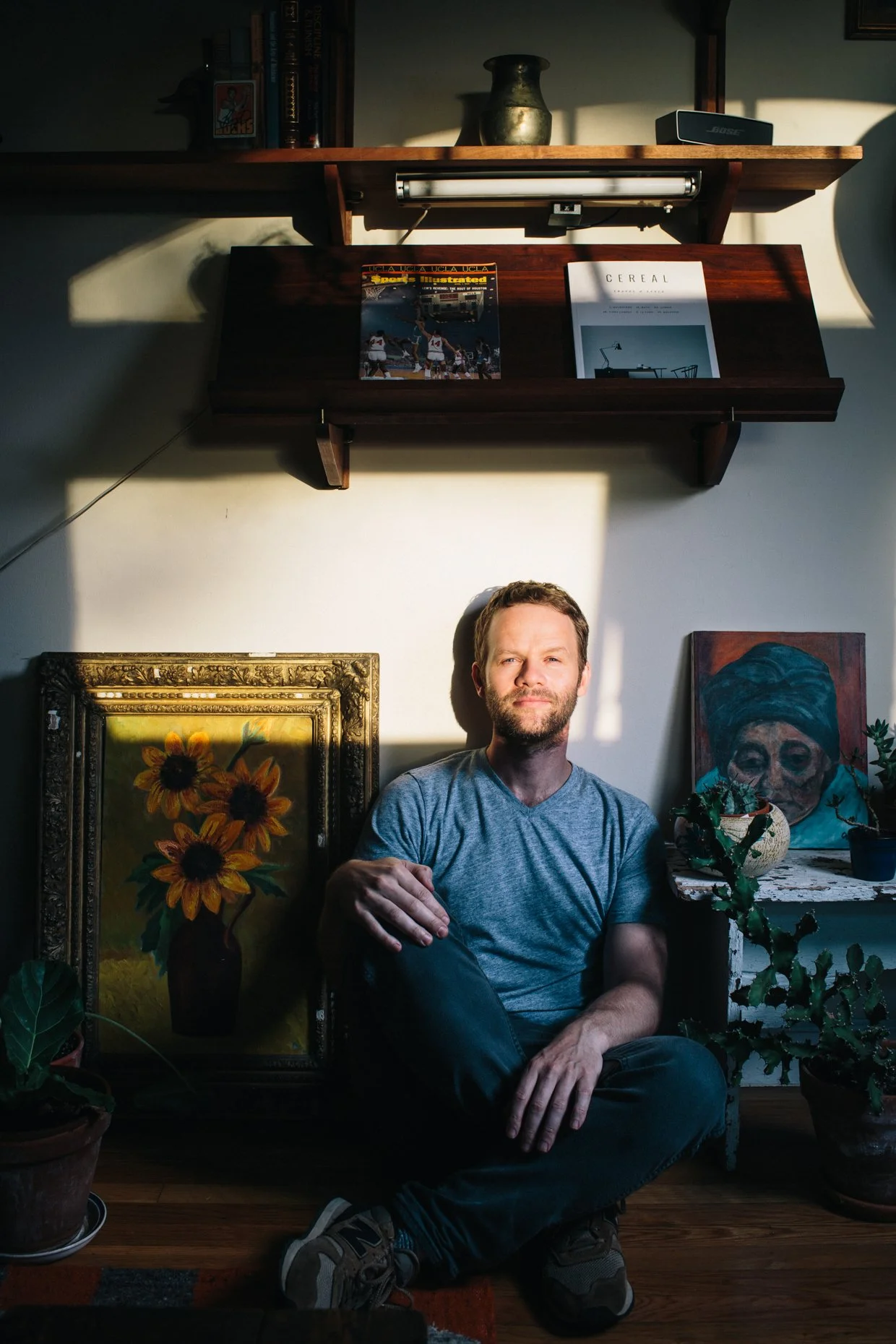 A man sitting in a room with sunlight casting shadows. Behind him are artworks, including a sunflower painting and a portrait of a woman wearing a headscarf. There are also potted plants on the floor.