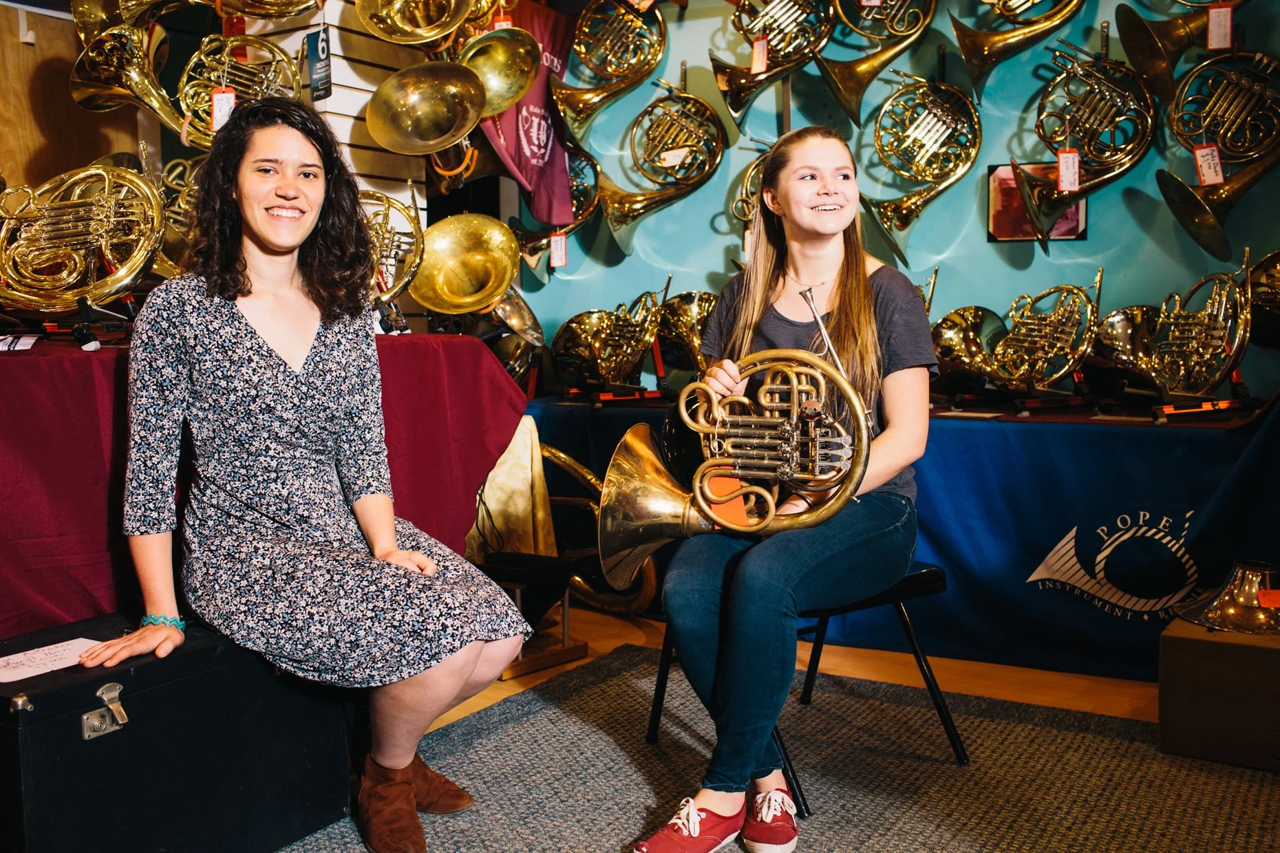 Two women in a music store, surrounded by brass French horns on display. One is sitting on a black case, smiling, and wearing a floral dress with brown boots. The other girl is seated, holding a French horn, smiling, and wearing a gray t-shirt, jeans